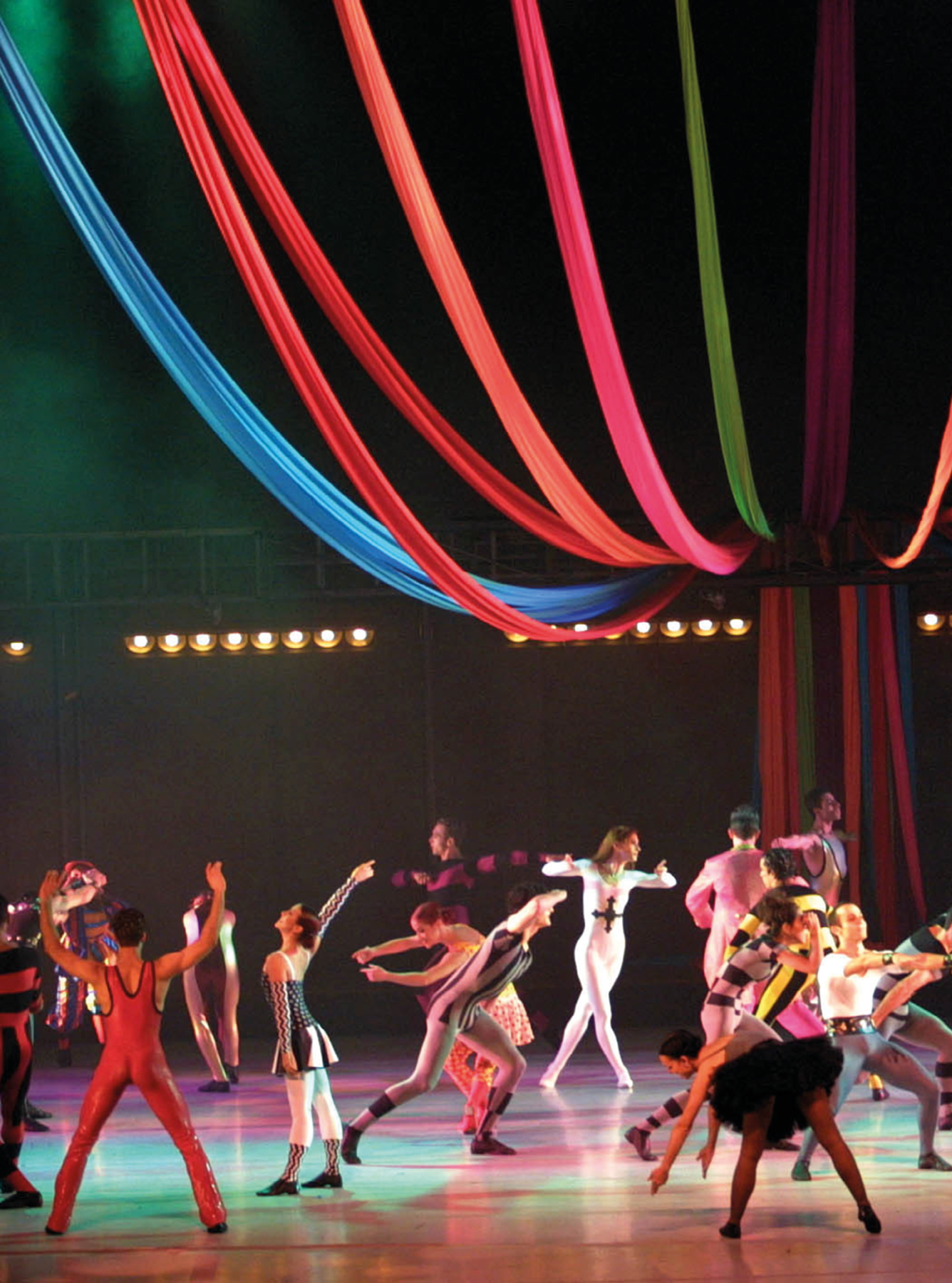 Fotografia. Diversos bailarinos com roupas circenses, distribuídos sobre um palco iluminado em várias cores. Todos estão em pé, mas em poses diferentes, fazendo uma performance. Na parte superior, tecidos coloridos suspensos formando ondulações que imitam a lona de um circo presa em um poste no centro do picadeiro.