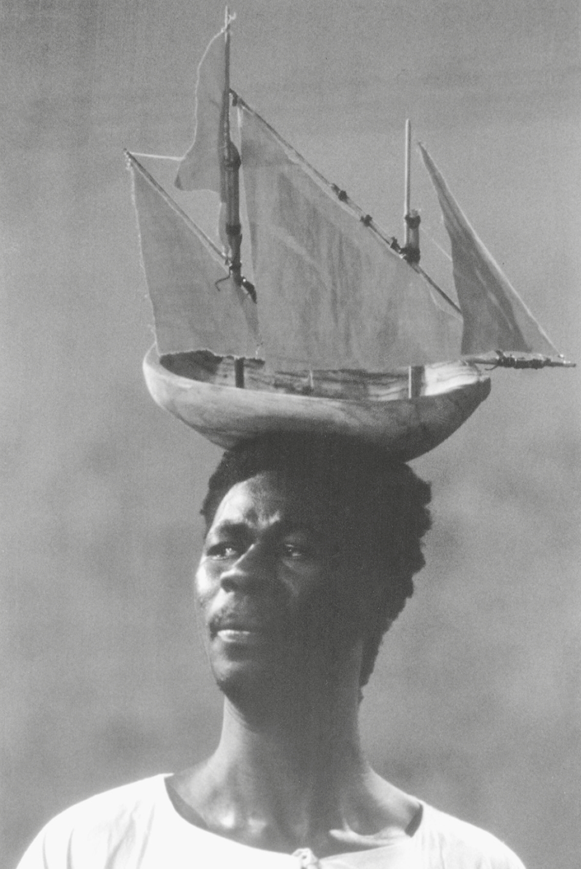 Fotografia em preto e branco. Retrato em meio perfil de um homem usando camiseta branca. Ele tem acima da cabeça, como um chapéu, um barco à vela, feito de madeira e tecido. Ele tem postura altiva e expressão séria.