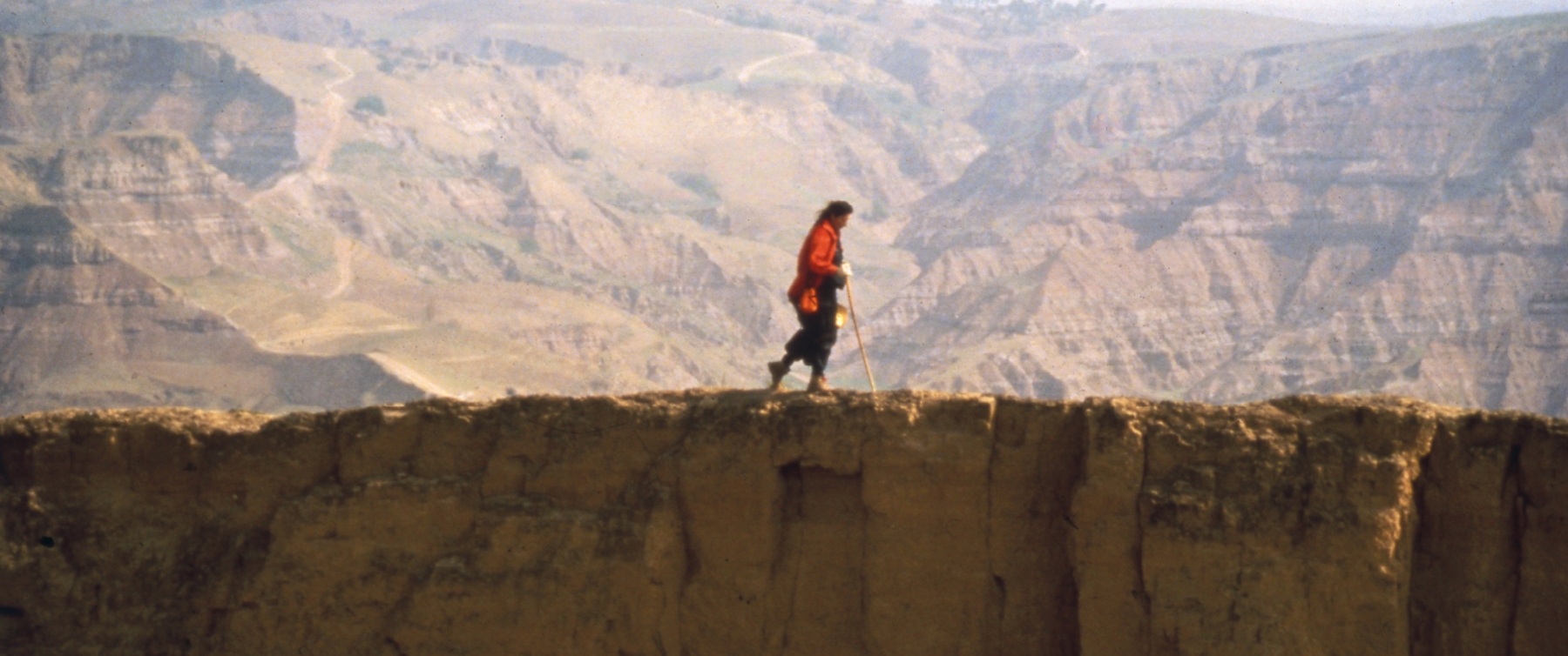 Fotografia. Pessoa de perfil, usando um casaco vermelho e trajes escuros, caminha apoiando-se em um bastão longo, no topo de uma muralha. Ao fundo, montanhas.