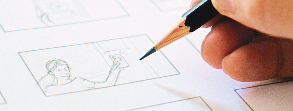 Fotografia. Destaque das pontas dos dedos de uma pessoa segurando um lápis preto sobre um quadrinho com ilustrações a traço.