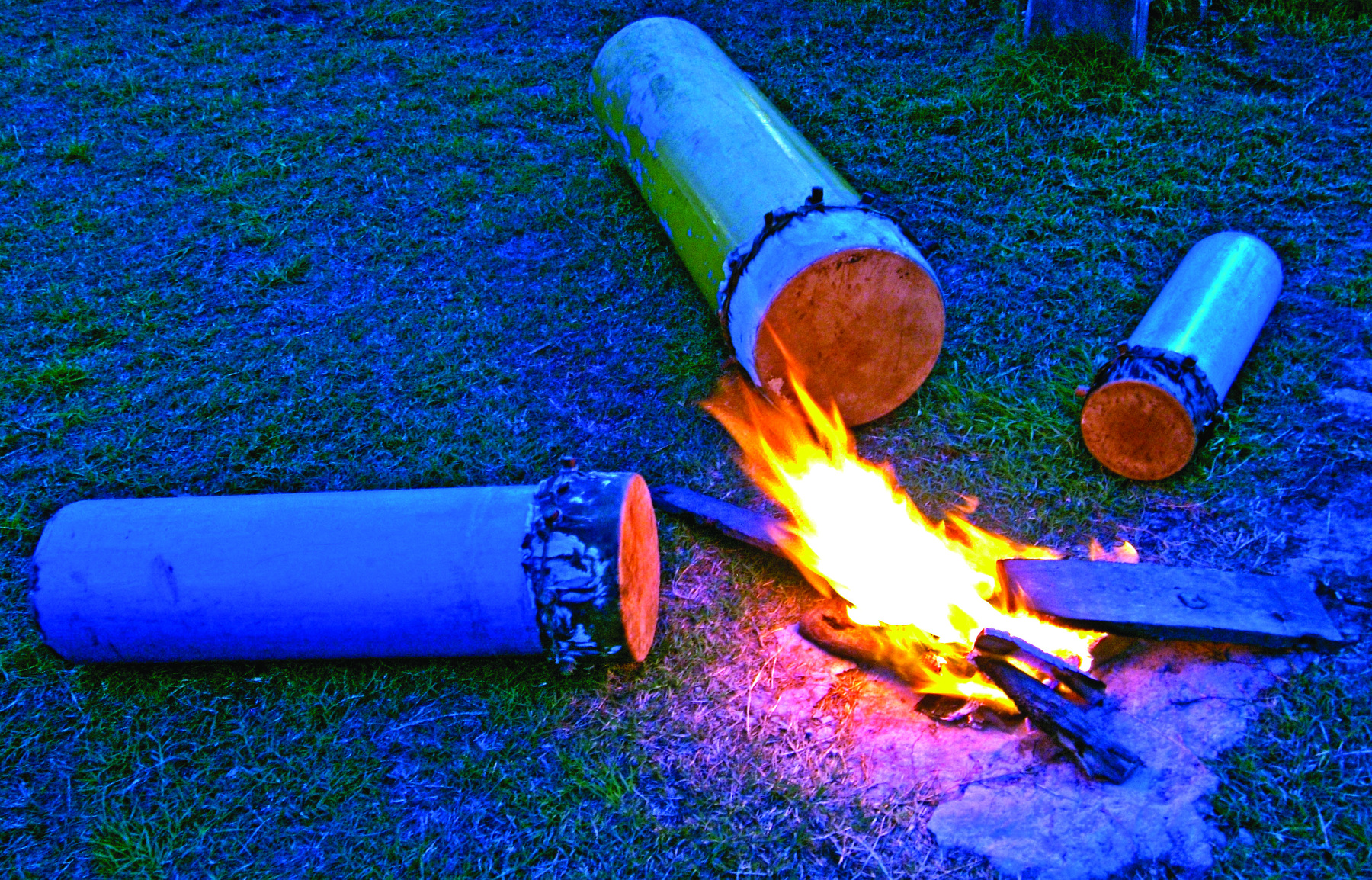 Fotografia. Três tambores de tamanhos diferentes ao redor de uma fogueira, sobre a grama.