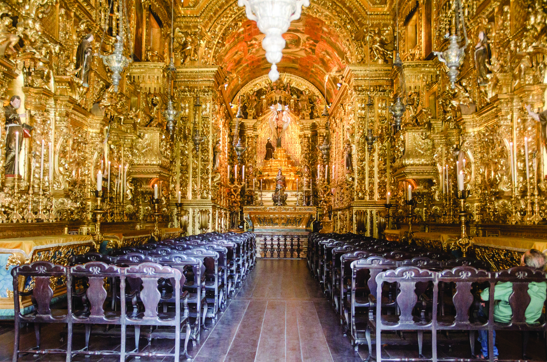Fotografia. Vista interna de igreja com paredes e colunas douradas com muitos detalhes. Nas laterais, há fileiras de cadeiras de madeira. Ao fundo, há um altar com uma escultura central.