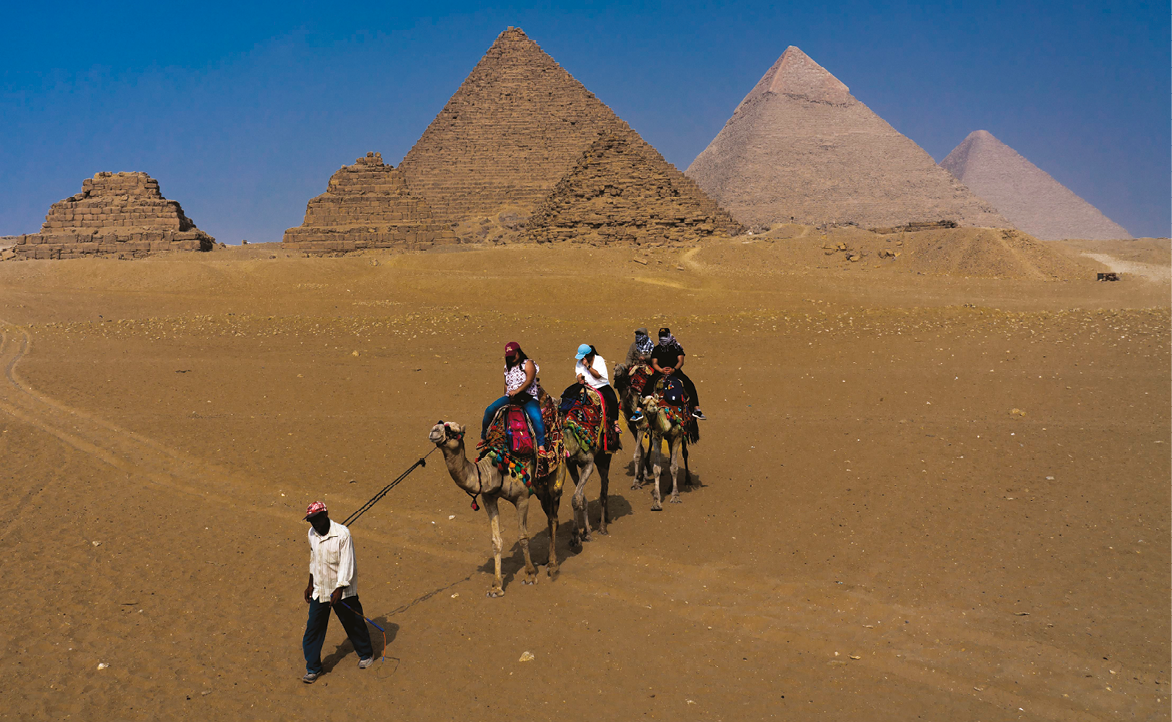 Fotografia. Um homem caminha em campo aberto segurando um cordão preso a um camelo. Sobre o camelo, há duas pessoas. Atrás, há duas pessoas montadas em outro camelo. Ao fundo, há três pirâmides altas e três pirâmides baixas. As duas da esquerda estão em ruínas.