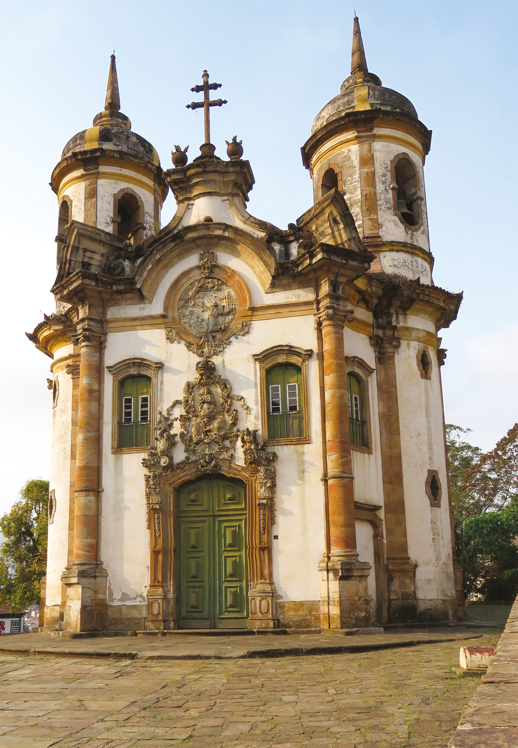 Fotografia. Fachada de  igreja com pintura branca desgastada e adornos dourados,  duas torres com sinos, uma cruz no centro, três pequenas janelas no alto e uma porta central verde.