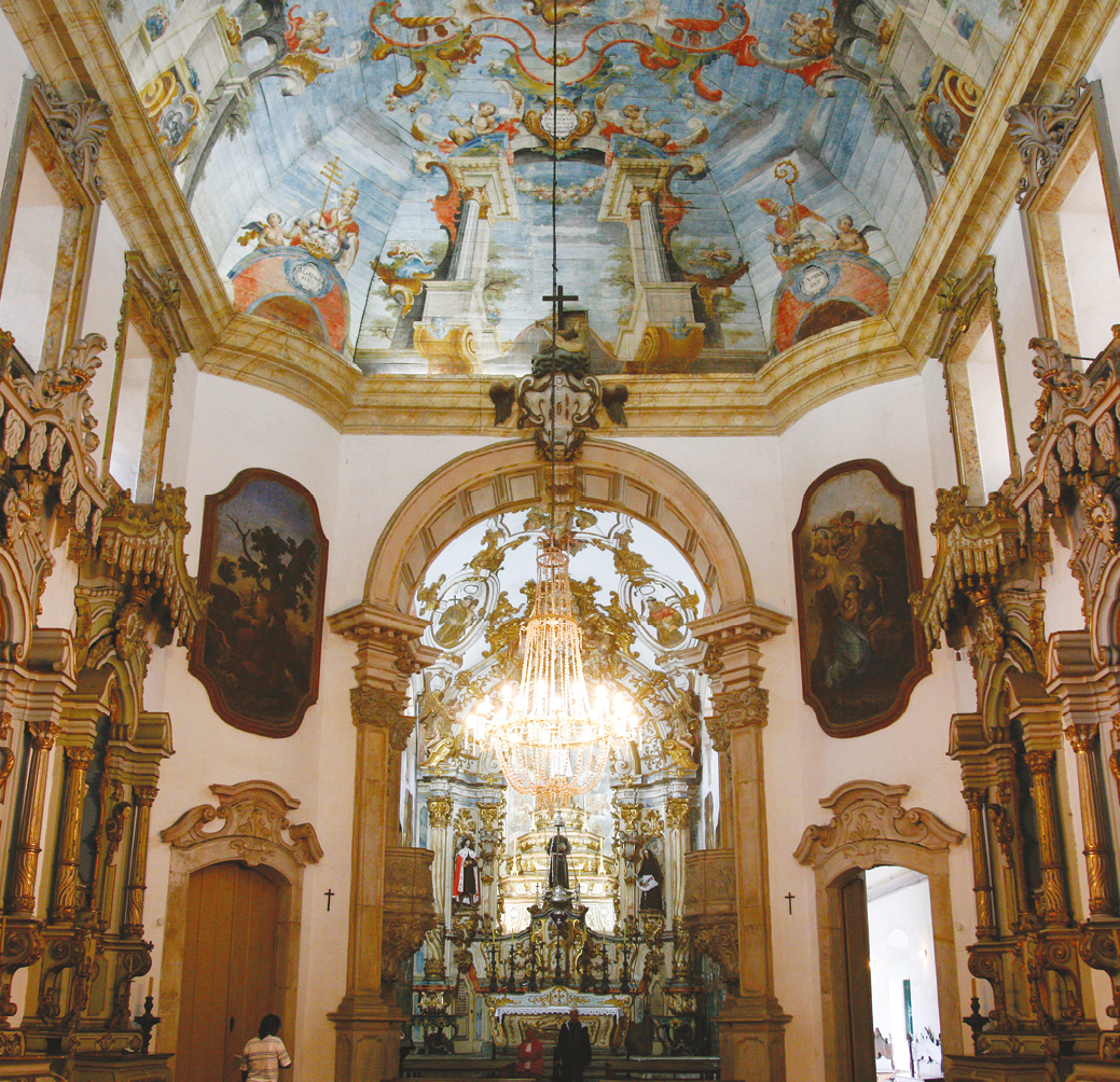 Fotografia. Vista interna de igreja. No centro, altar com pinturas azuis e douradas. Nas laterais, há pilares dourados. No teto, pintura de figuras angelicais com predomínio das cores azul, vermelho e dourado.