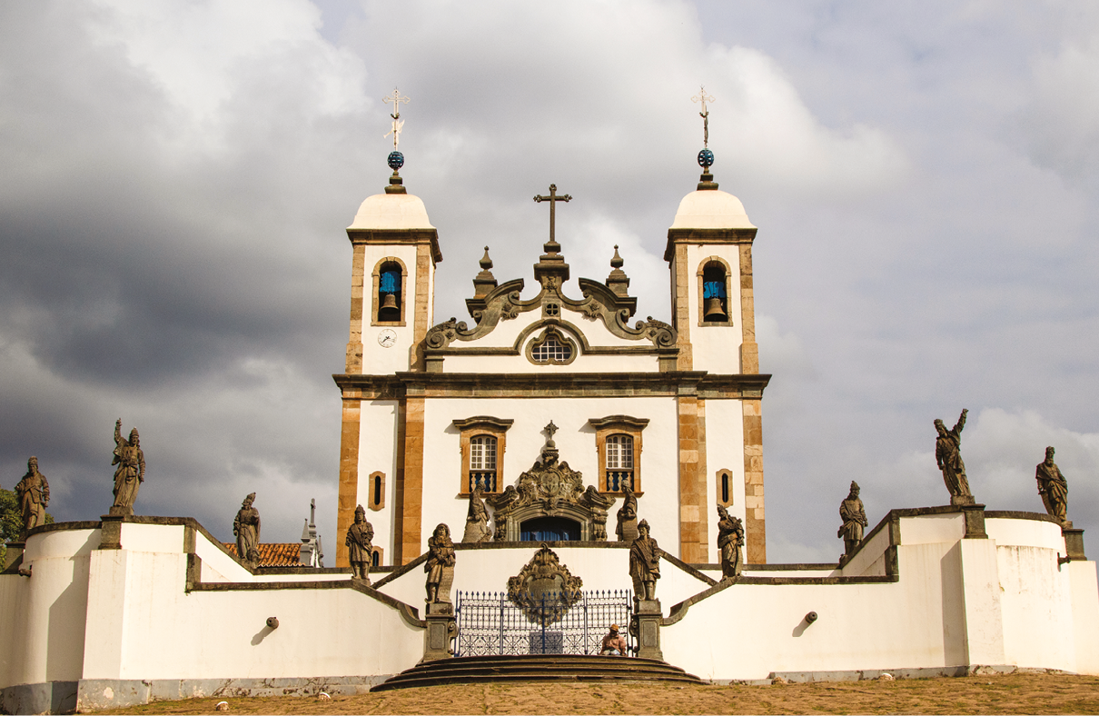 Fotografia. Fachada de uma igreja com paredes brancas e detalhes na cor bronze com duas torres com sino. No centro, na parte superior, há uma cruz. Na frente, escadaria com esculturas.