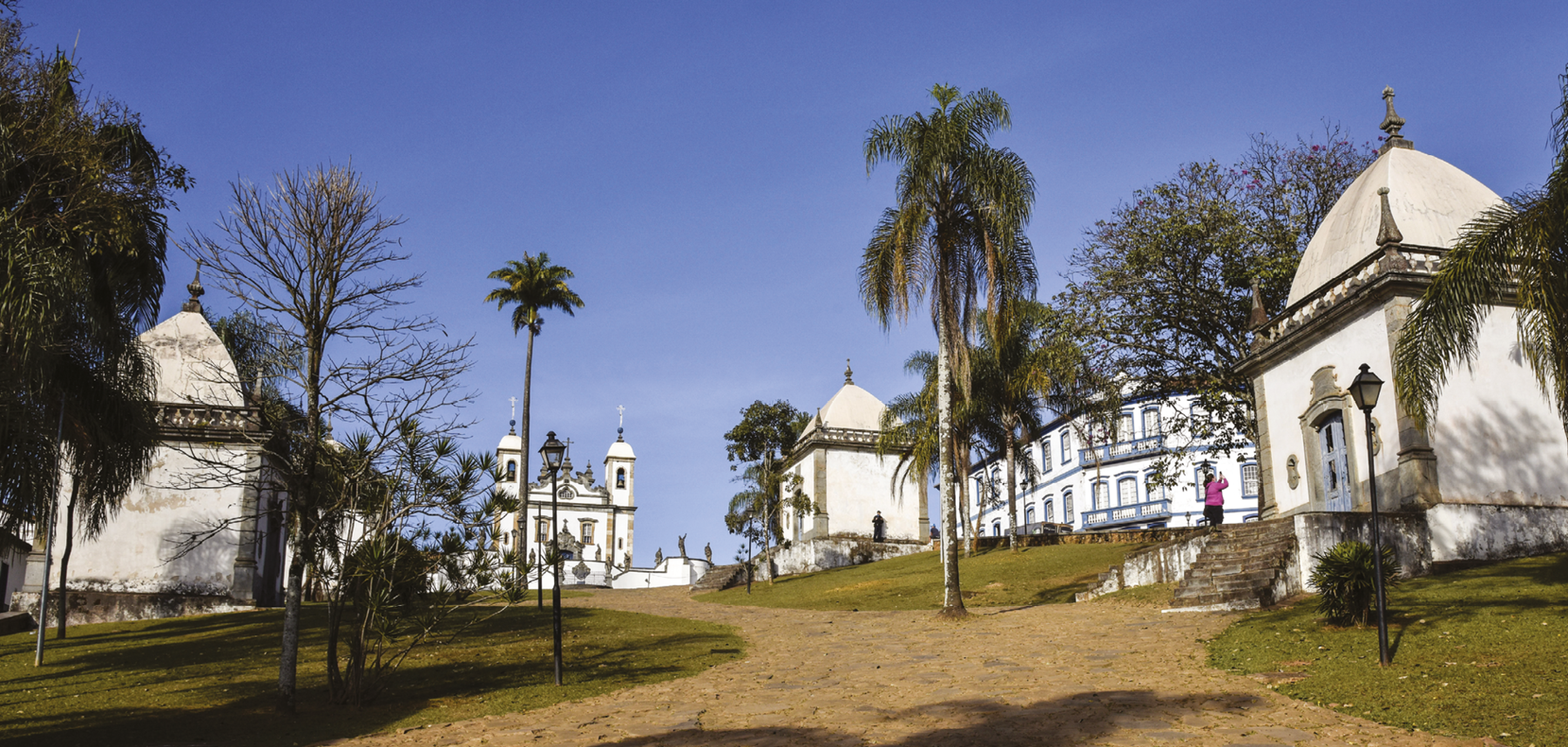 Fotografia. Pátio aberto com grama nas laterais, árvores esparsas, arandelas de jardim e um caminho de pedras. Nas laterais, há duas capelas. Ao fundo, há uma igreja pintada de branco e detalhes na cor bronze com duas torres com sino. No centro dela, na parte superior, uma cruz. Na frente, escadaria com esculturas. Do lado direito, atrás, há uma construção azul e branca.