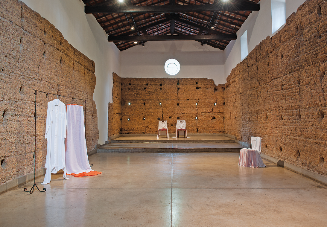 Fotografia. Uma sala, com tijolos nas paredes e pintura branca acima dos tijolos, telhas e vigas de madeira aparentes no teto e chão de cimento queimado cinza. À esquerda, uma arara de roupas com uma túnica branca. Ao lado, um tecido branco e laranja pendurado. À direita, uma cadeira branca. Ao fundo, duas cadeiras brancas.