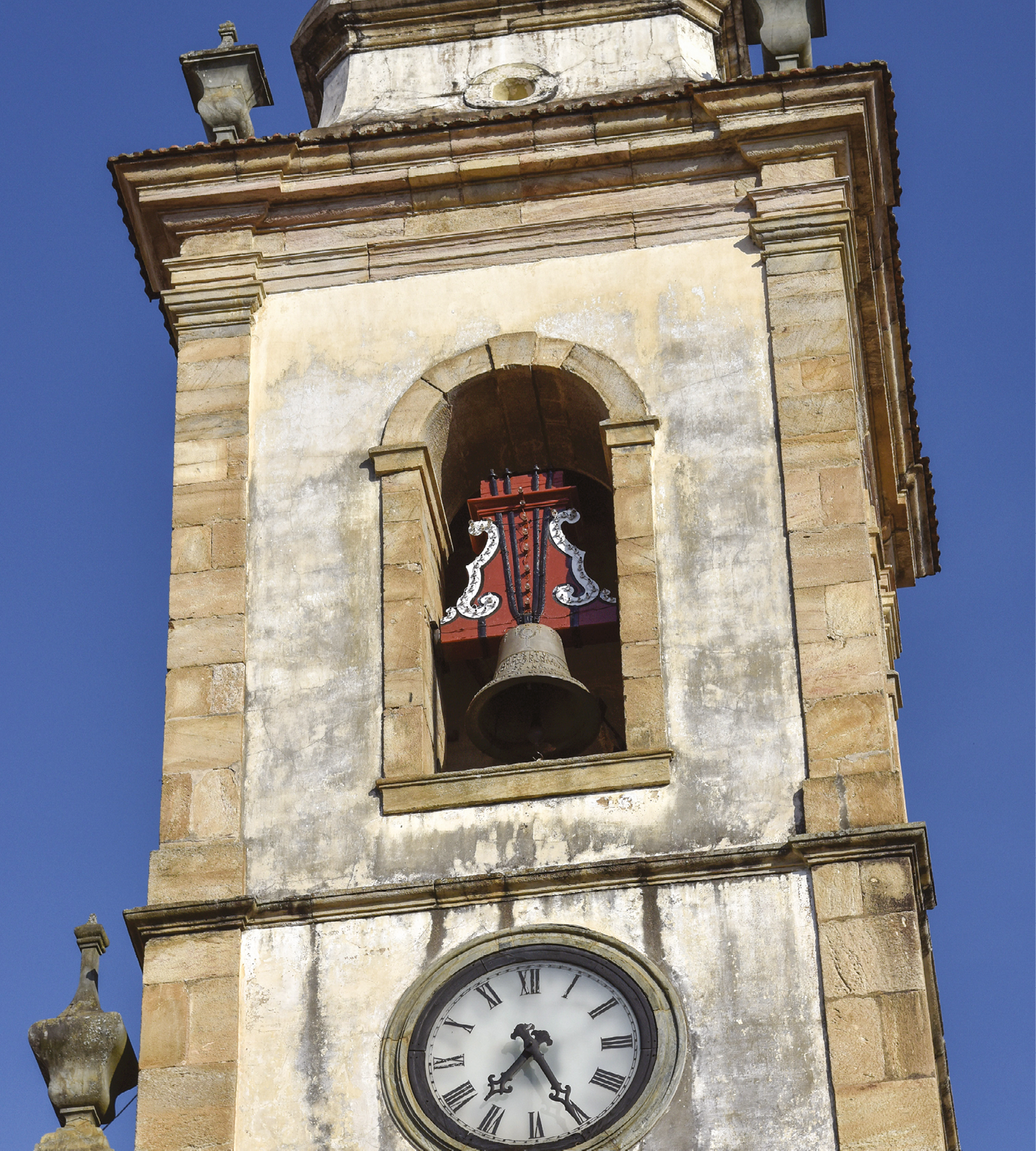 Fotografia. Destacando uma torre com um sino. Abaixo, um relógio com números romanos.