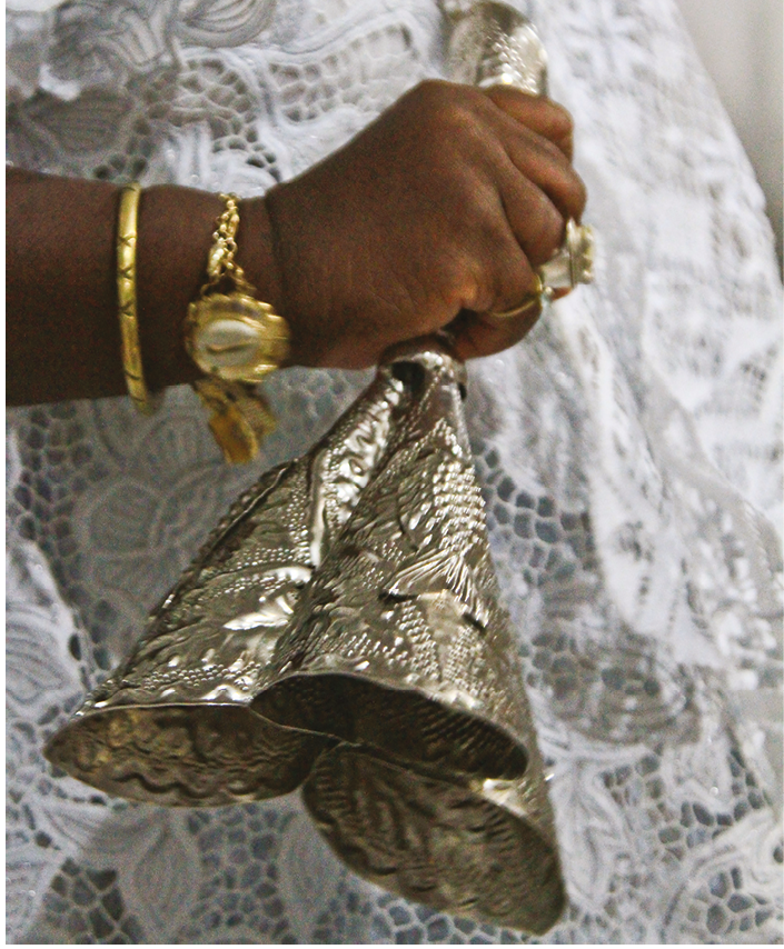 Fotografia. Destaque para uma mão negra com pulseiras  e anéis dourados  um adjá (instrumento com três sinos de pratas). Atrás, renda branca de um vestido.