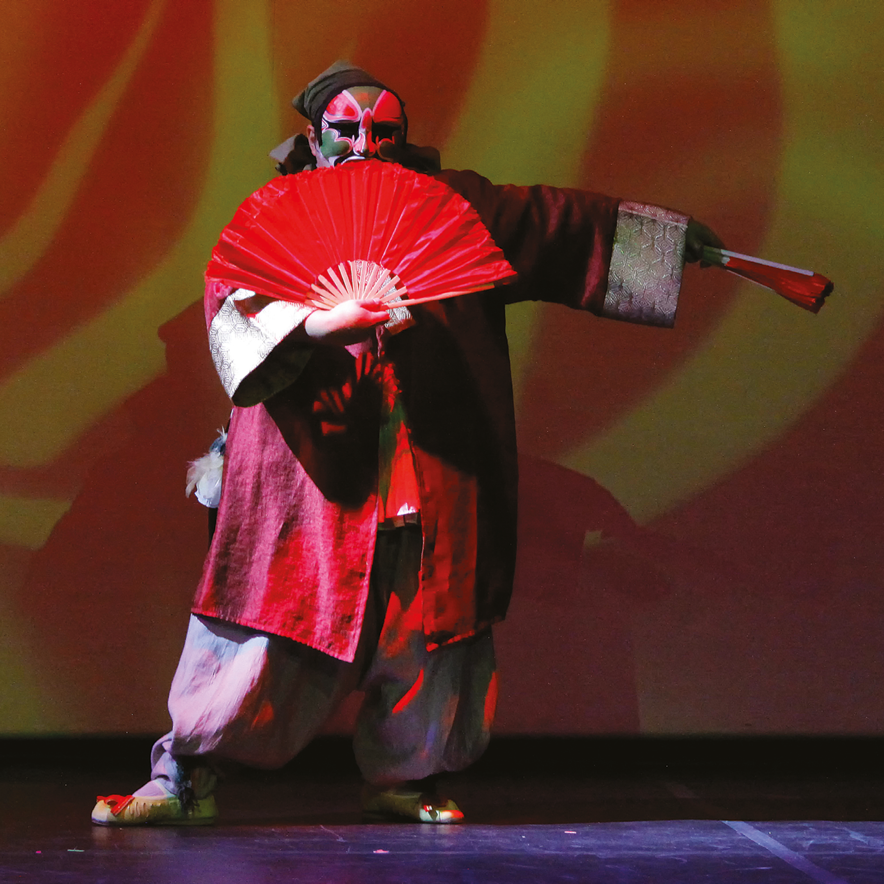 Fotografia. Uma pessoa com espécie de kimono vermelho de punhos prateados, calça larga clara, sapatilhas e máscara pintada de vermelho e verde no rosto. Ele está de pé e segura um leque vermelho aberto na frente do corpo com a mão direita e um leque fechado com a mão esquerda. O braço esquerdo está estendido na lateral do corpo. No fundo, há sombras e luzes vermelhas e amarelas.