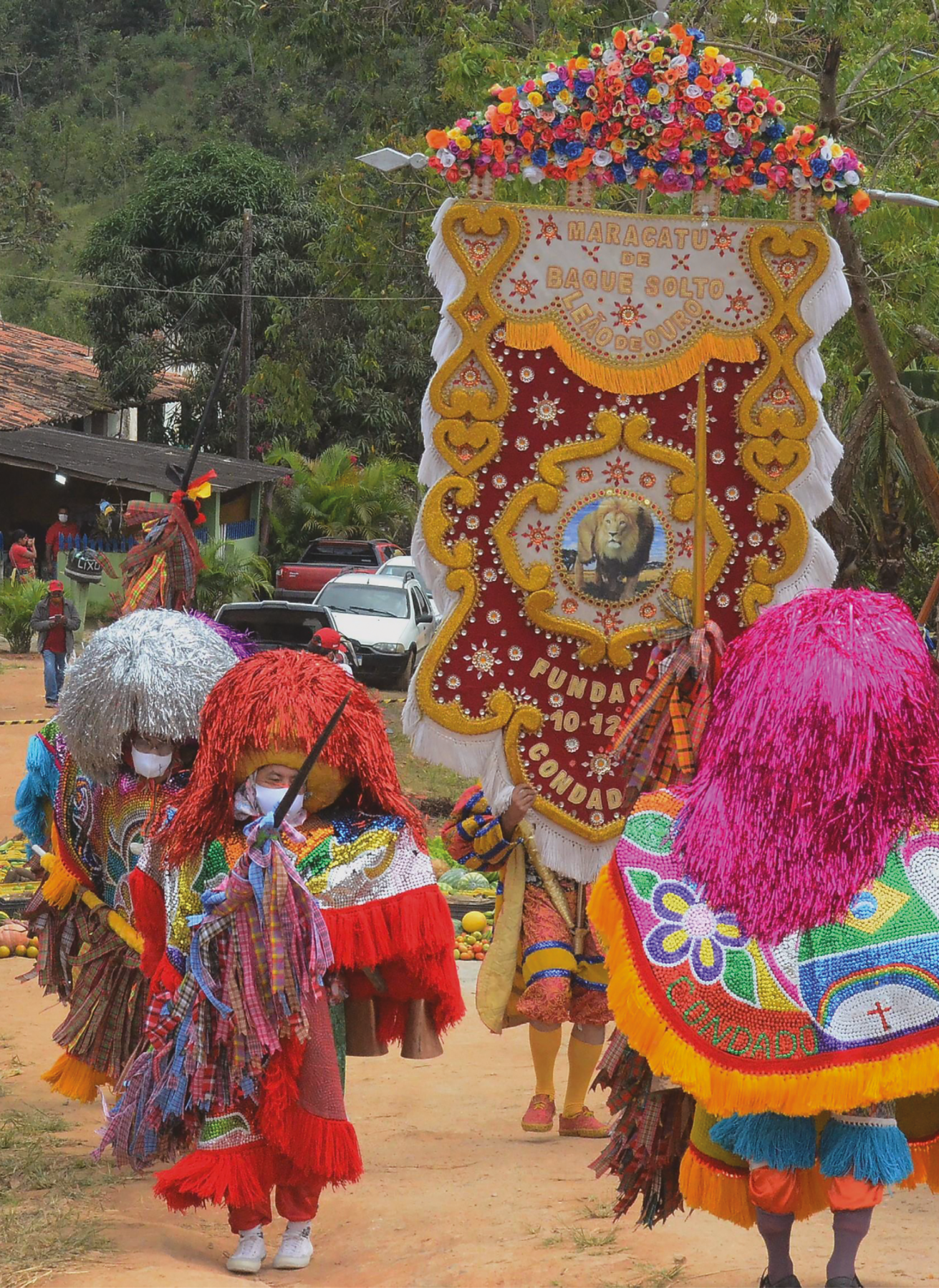 Fotografia. Três pessoas enfeitadas usam chapéu com peruca colorida e brilhosa, ponchos coloridos com desenhos da bandeira do Brasil e de Pernambuco e de flores. Atrás, uma pessoa caminha e segura um estandarte vermelho, amarelo e dourado do Condado, composto de flores e do nome na parte de cima, em letras douradas e fundo branco com flores, MARACATU DE BAQUE SOLTO - LEÃO DE OURO; ao centro do estandarte, fotografia de um leão; na parte inferior do estandarte, data de fundação e nome nas cores vermelha e dourada. Ao fundo da imagem, há pessoas em casas simples e carros estacionados em rua de terra. Atrás, há árvores.