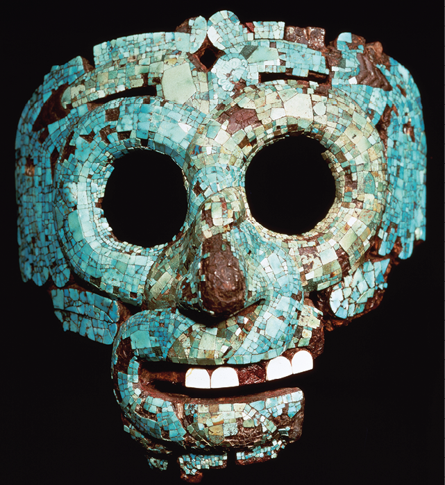 Fotografia. Máscara de um crânio, azul com detalhes em marrom. Os olhos são dois círculos vazados, sem preenchimento, o nariz é proeminente e na boca semiaberta há quatro dentes brancos, intercalados por espaços em marrom.