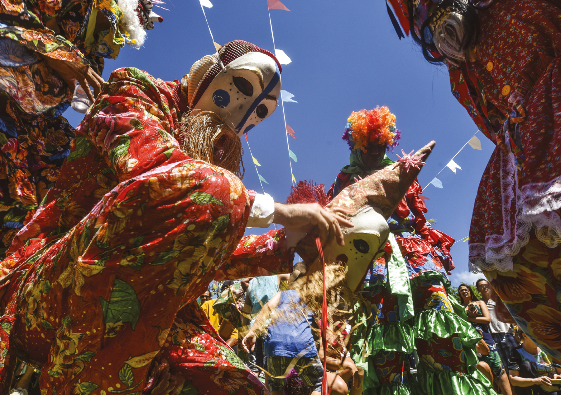 Fotografia. Apresentação ao ar livre. Em primeiro plano, à esquerda, uma pessoa com roupa colorida, predominante vermelha com floral amarelo e verde, e máscara branca com detalhes de pintura em azul, com palha pendurada que sai da boca. Ela está agachada, com o joelho esquerdo apoiado no chão e segura nas mãos uma máscara branca com detalhes em vermelho. No centro, ao fundo, uma pessoa em pé, com roupa colorida predominantemente vermelha com floral azul, com camadas de babados verdes, peruca colorida cor de laranja e vermelha, olha para o chão. Ao redor, outras pessoas com figurinos semelhantes. Atrás, pessoas sem fantasia observam a cena. Acima, e bandeirinhas penduradas em fios sob céu azul.