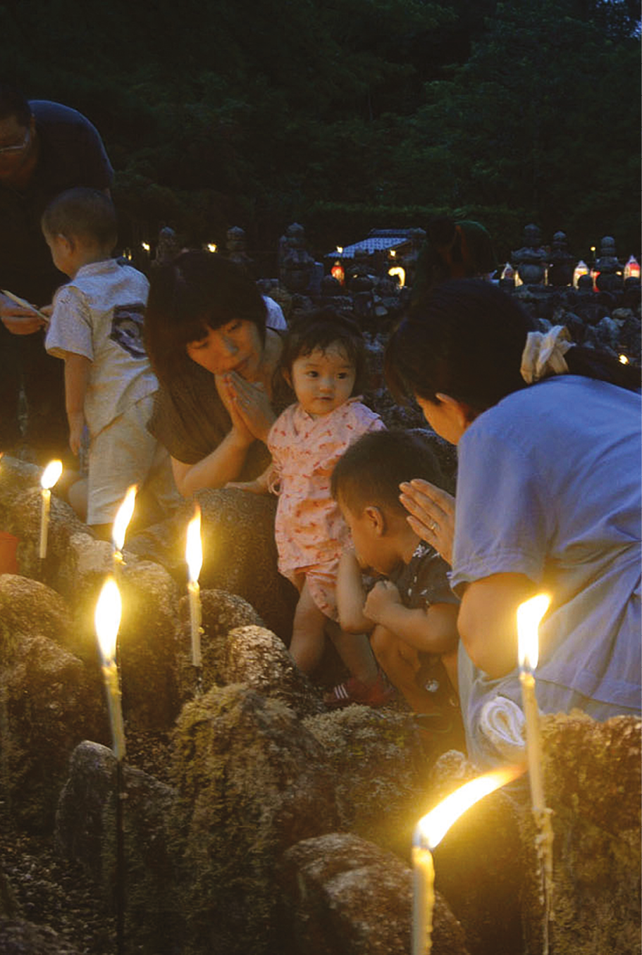 Fotografia. Em primeiro plano, algumas velas acesas em pedras, ao ar livre. À esquerda, um homem passa um objeto para uma criança vestida de branco. Ao lado, uma mulher com as mãos em oração olha para uma menina pequena que está ao lado dela. Ao lado da menina, um menino agachado diante das pedras. À direita, uma mulher vestida de azul, de costas, as mãos em oração, olha para as crianças. Ao fundo, aparecem túmulos.