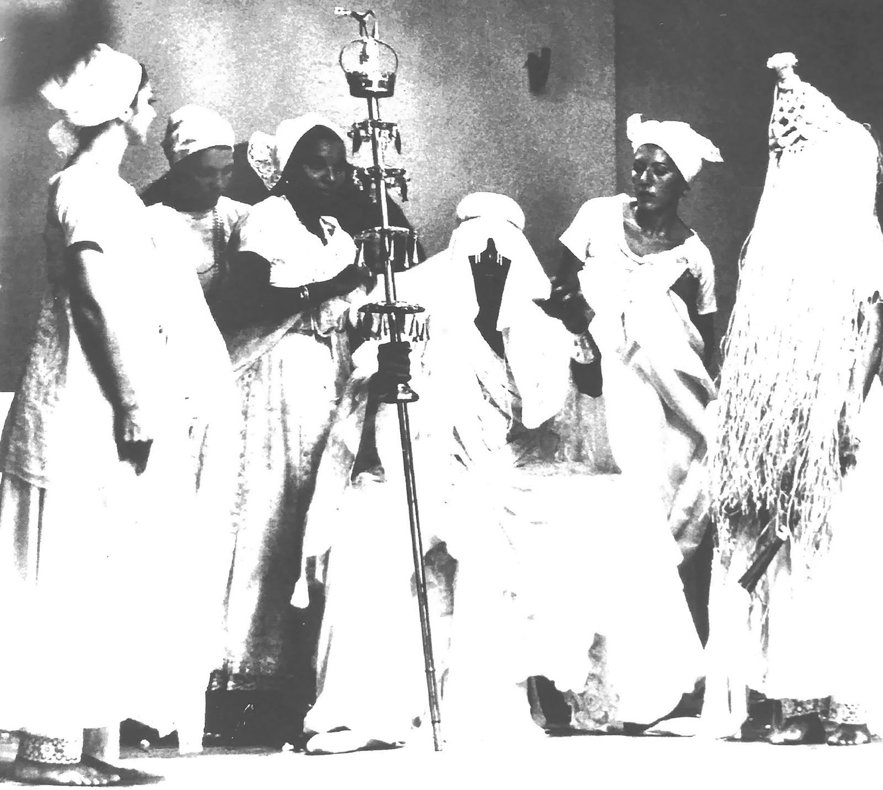 Fotografia em preto e branco. Espetáculo. Mulheres usam lenço na cabeça e vestido brancos. À direita, uma pessoa está com o rosto coberto por palha. Ao centro uma pessoa segura um cetro de metal.