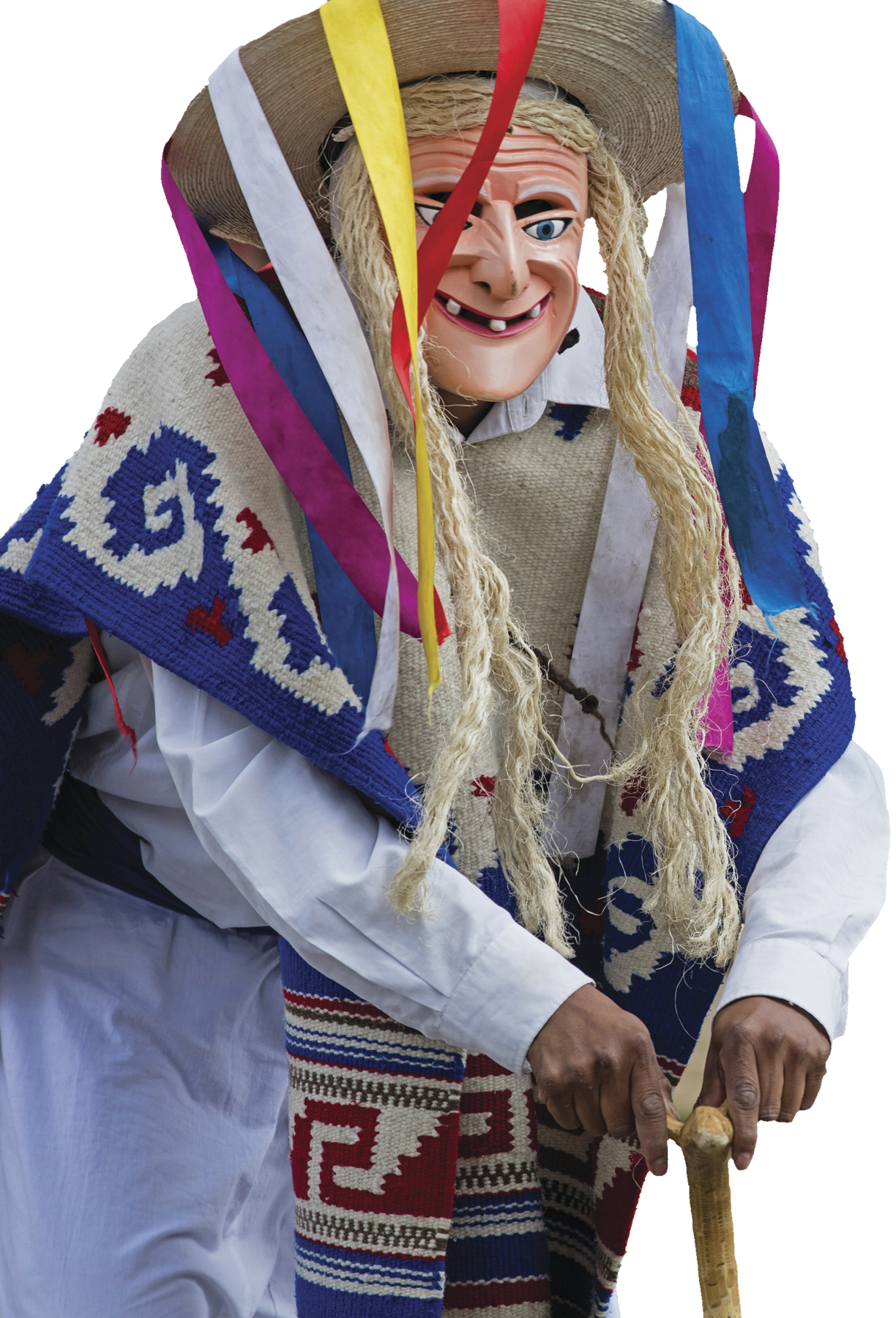 Fotografia. Detalhe de pessoa usando roupas brancas, uma máscara de idosa, com cabelos loiros feitos de palha e chapéu de palha com fitas coloridas que saem do topo do chapéu, e manto colorido. Ela está apoiada em uma bengala de madeira com as duas mãos.
