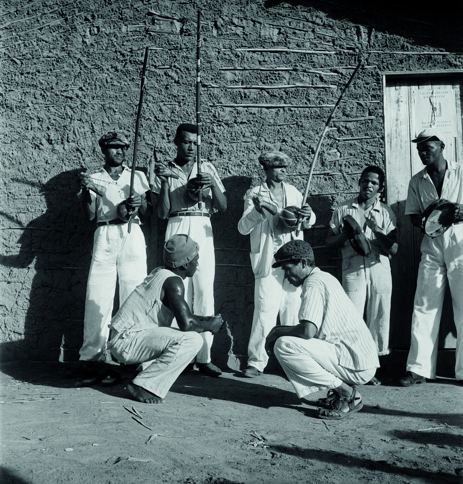 Fotografia em preto e branco. Em primeiro plano, à esquerda, dois homens frente a frente . Eles usam calça e camisa  claras e gorro. Ambos estão de perfil e agachados em um chão de terra.  Em segundo plano, à esquerda,  três homens usam roupas claras e seguram nas mãos um berimbau cada um. à direita, outros  dois homens com roupas claras e boné seguram um pandeiro cada um. Ao fundo, parede de casa de pau a pique.