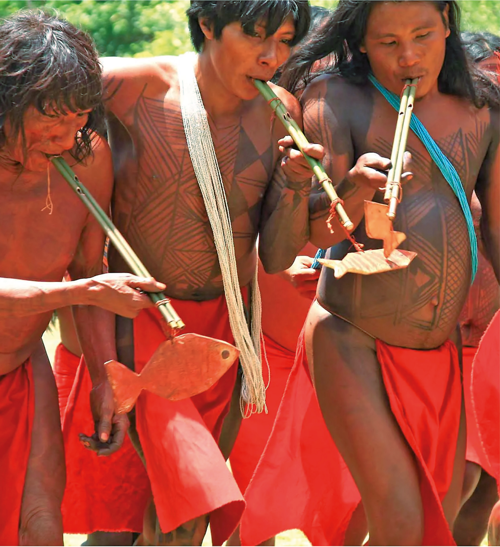 Fotografia. Três homens indígenas vestem uma espécie de tanga vermelha e têm o tronco pintado.  Eles tocam com a boca um instrumento de feito de bambu.