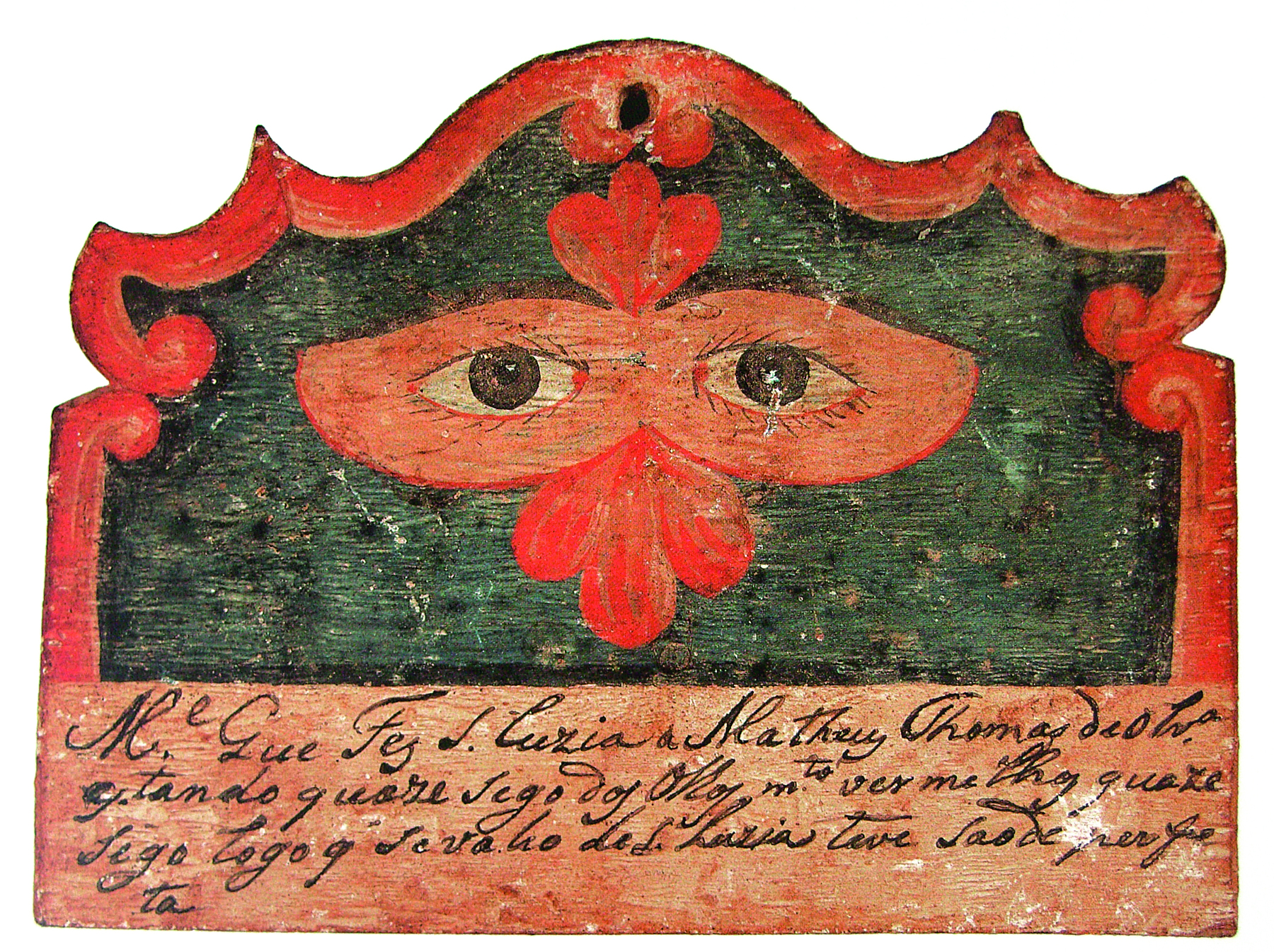 Fotografia. Escultura de madeira nas cores vermelho e preta. Na parte superior, ao centro, desenho de olhos castanhos, envoltos em um desenho em formato de máscara de olhos vermelha. Na parte inferior, texto escrito em letra cursiva.