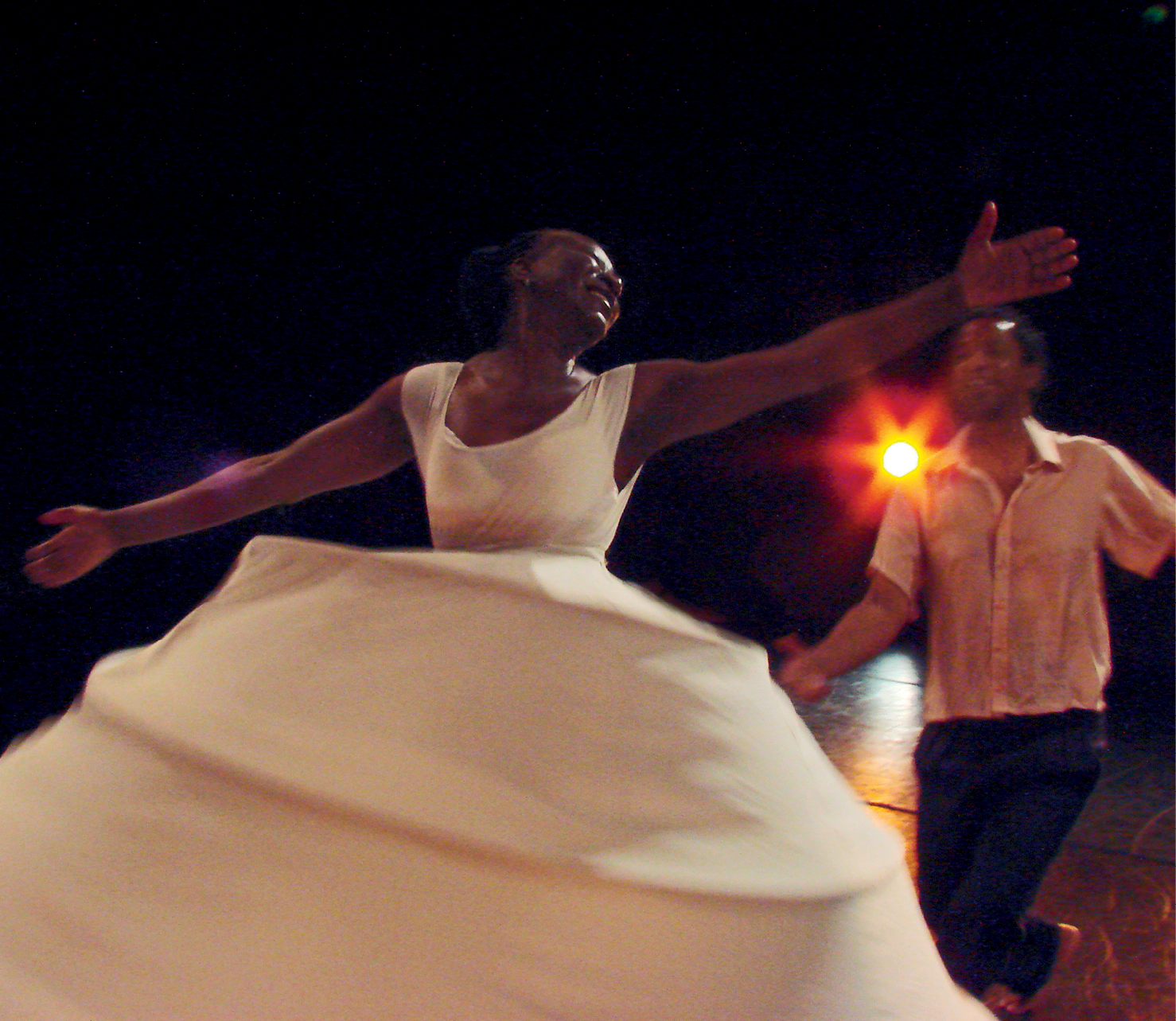 Fotografia. Espetáculo de dança. Mulher negra usa vestido branco rodado. Ela está em um giro com os braços abertos e sorri. Ao lado dela, um homem usa camisa branca e calça escura. Ele está com a perna direita flexionada com o pé para trás e o braço direito estendido na direção da mulher. Ao fundo luz laranja entre os dois.