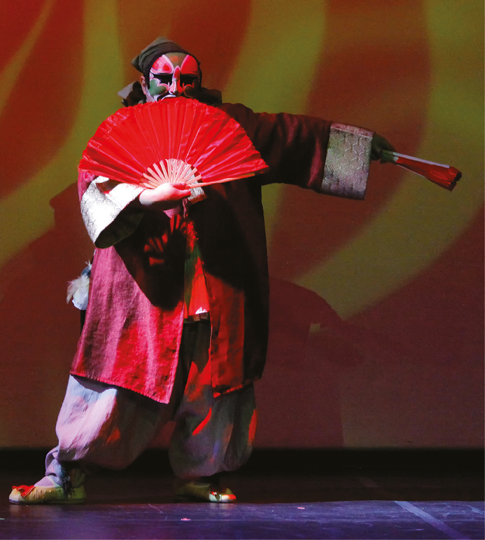 Fotografia. Uma pessoa com espécie de kimono vermelho de punhos prateados, calça larga clara, sapatilhas e máscara pintada de vermelho e verde no rosto. Ele está de pé e segura um leque vermelho aberto na frente do corpo com a mão direita e um leque fechado com a mão esquerda. O braço esquerdo está estendido na lateral do corpo. No fundo, há sombras e luzes vermelhas e amarelas.