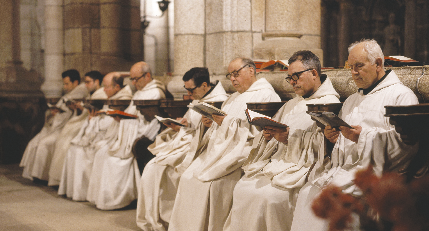 Fotografia. Sete homens de cabelos curtos, alguns escuros outros grisalhos,  usando túnicas brancas. Alguns usam óculos. Eles estão sentados em fileira, lado a lado, e seguram com as mãos um livro aberto. Ao fundo, pilares e paredes de mármore.