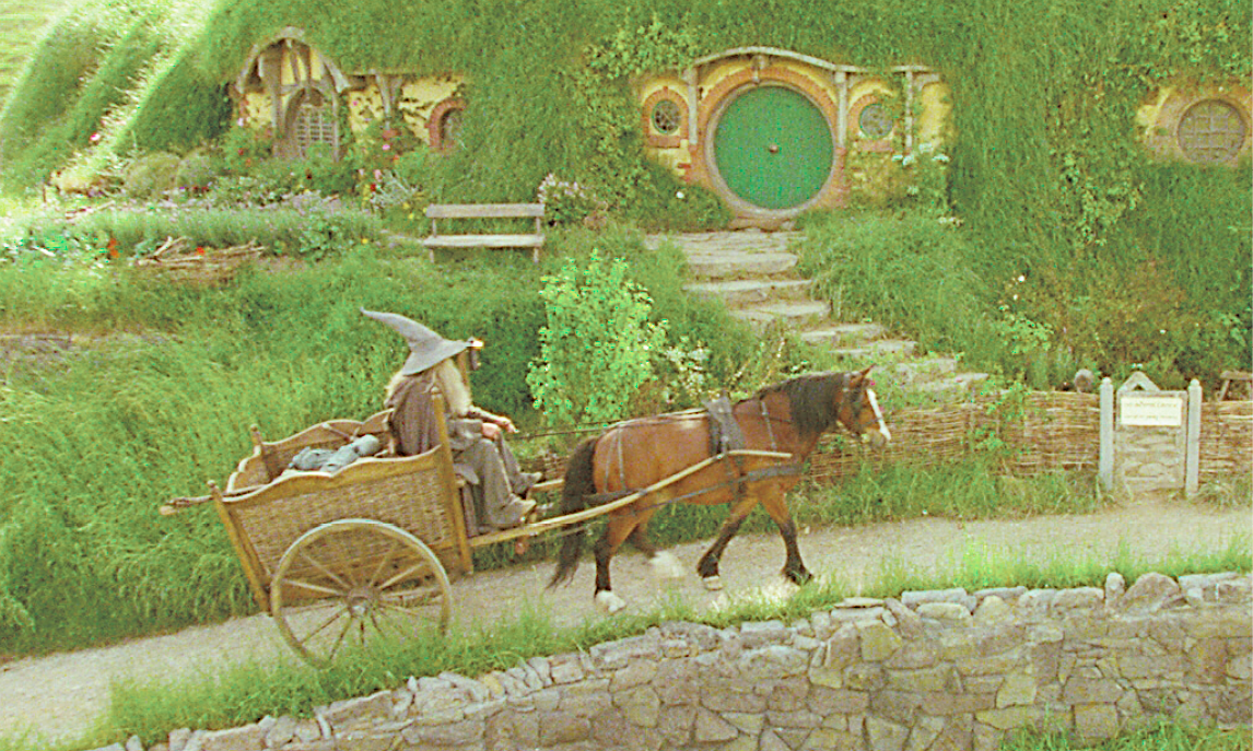 Fotograma.  Cena de filme. Um homem de chapéu pontudo e roupas cinzas está sentado em uma carroça guiado por  um cavalo marrom, que percorre um caminho estreito. Do lado esquerdo do homem, há uma escada de pedra que leva a uma  casa nas raízes de uma árvore envolta em grama.