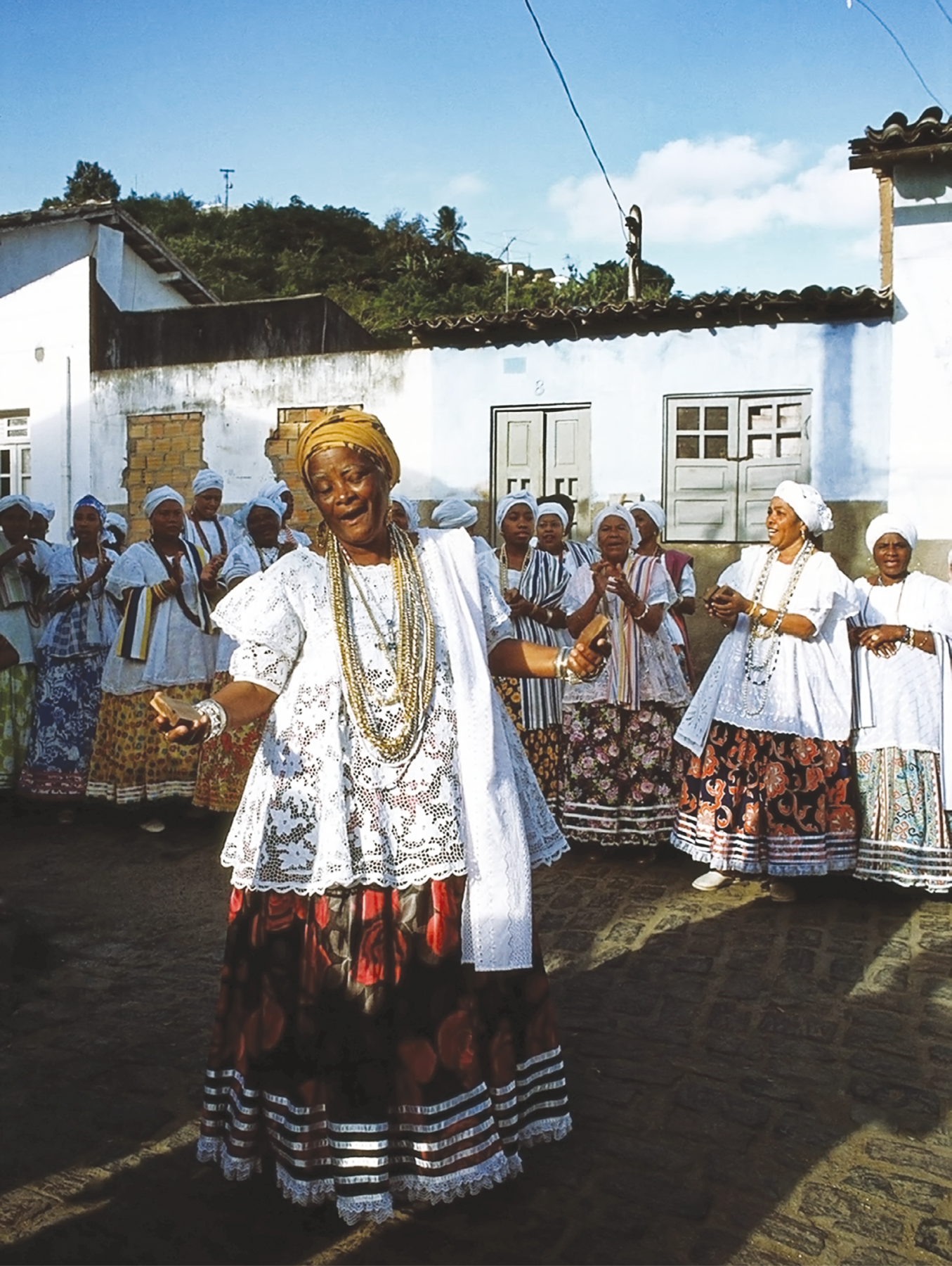 Fotografia. Em primeiro plano, ao centro, uma mulher de meia idade, usando saia rodada e florida comprida, bata de renda branca, colares no pescoço e um turbante na cabeça está de pé com os olhos fechados e os braços flexionados para a frente, com a boca aberta. Ela parece dançar e cantar e segura um pedaço de madeira em cada  mão. Atrás dela, outras mulheres usam roupas semelhantes. Algumas batem palmas. Todas estão em uma rua de pedra. Ao fundo, construção de parede branca e janelas cinza.