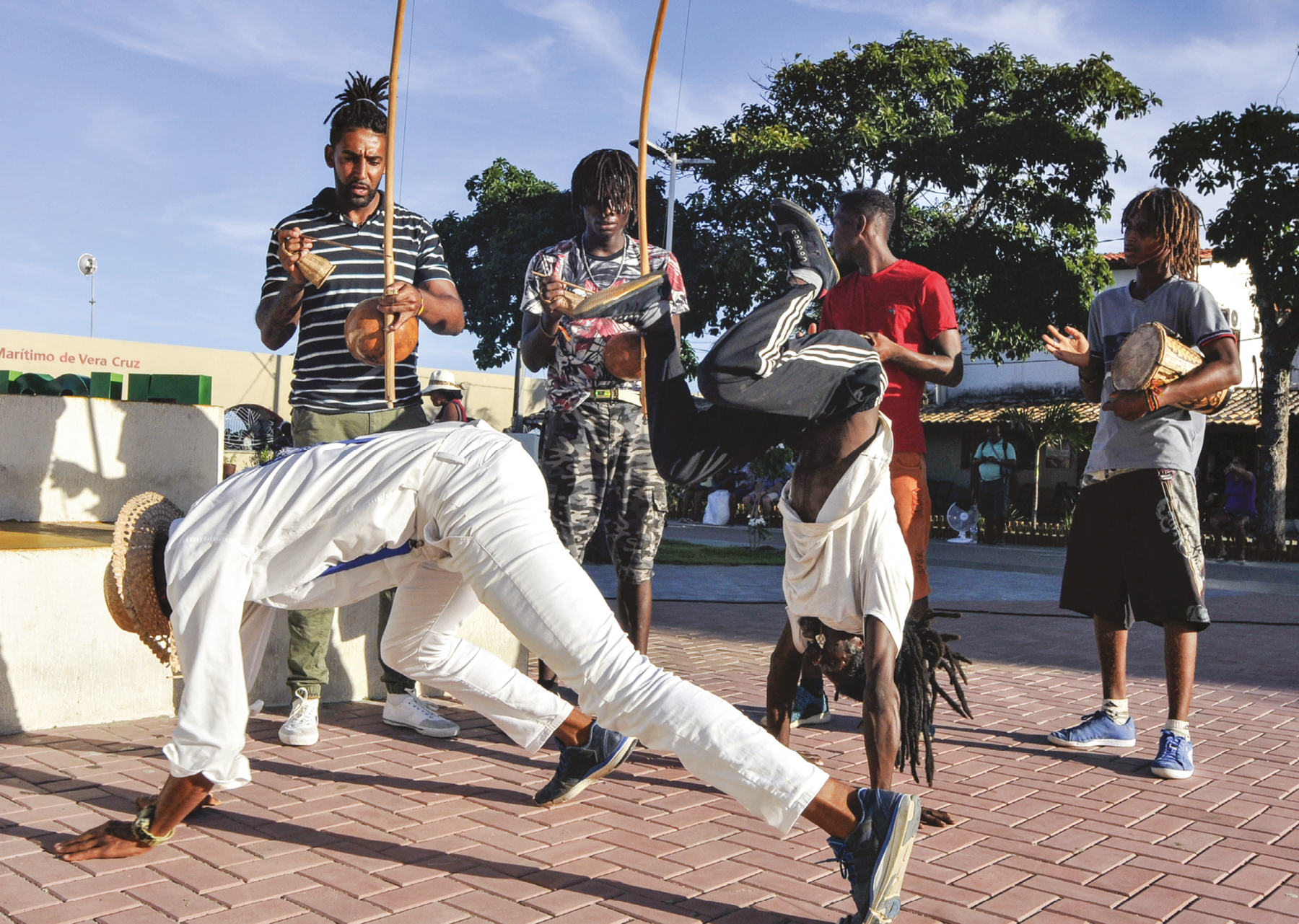 Fotografia. Em primeiro plano,  um homem de roupas brancas, chapéu e tênis está com as mãos e os pés no chão e o tronco elevado, jogando capoeira. Ao lado dele, um menino de camiseta branca e calça preta está de ponta-cabeça com as mãos no chão e as pernas dobradas ao ar. Atrás, dois homens tocam berimbaus e ao lado deles, outros dois homens tocam tambores. Todos estão em uma calçada. Ao fundo, árvores e casas.