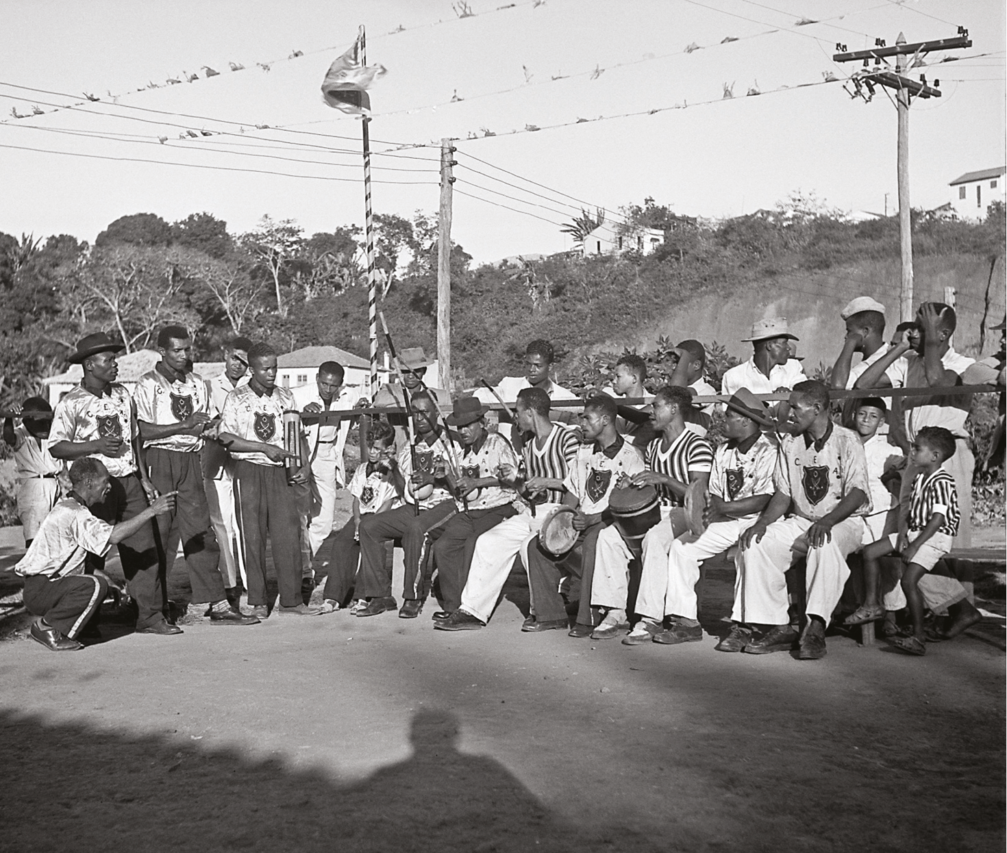 Fotografia em preto e branco. À esquerda, um homem usa camiseta branca e calça listrada e está agachado, de perfil, com o braço direito estendido para a frente, apontando o dedo indicador. Ao lado, um grupo de homens segura instrumentos musicais - berimbau, tambor e pandeiro. Alguns estão em pé e outros, sentados. Atrás, um grupo de homens, adolescentes e meninos observa a cena; alguns em pé, apoiados com os cotovelos em uma barra de madeira, outros sentados em banco de madeira. Ao fundo, árvores.