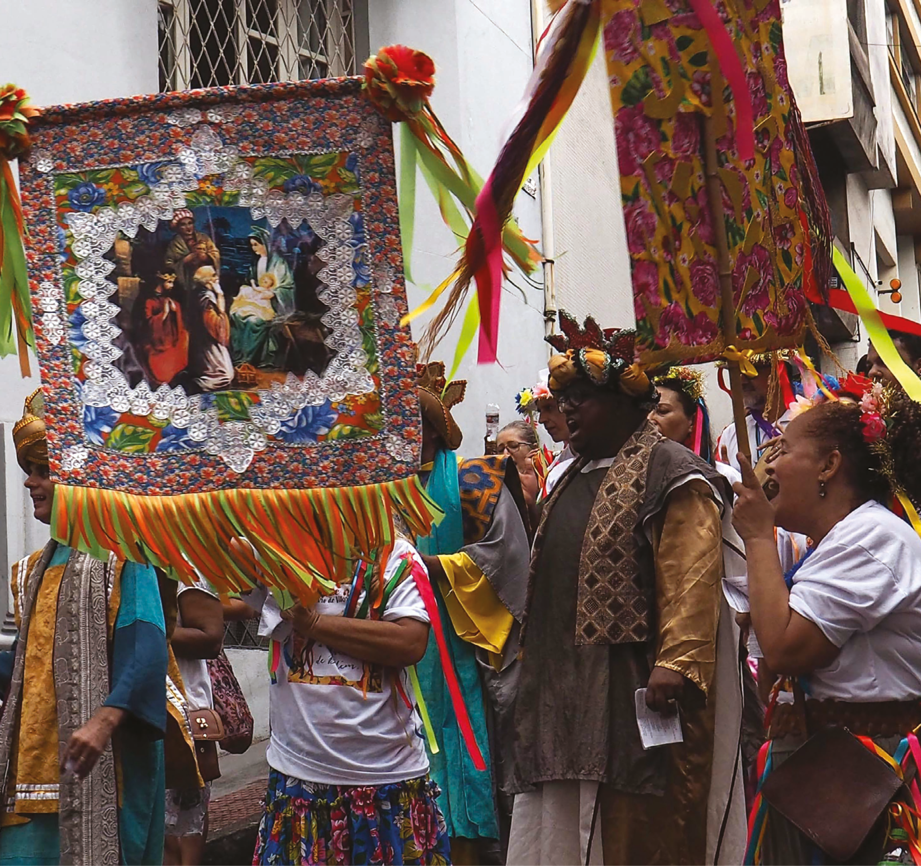 Fotografia. Grupo de pessoas caminhando. À esquerda, um homem de turbante dourado e túnica predominantemente azul. Ao lado dele, uma pessoa com camiseta branca e saia de chita estampada segura com as mãos, à frente do rosto, um estandarte com a pintura de Maria e Jesus em seu colo. No centro da imagem, um homem de óculos, com coroa e túnica marrom, com echarpe em tons de marrom e mangas douradas, está de boca aberta e segurando um papel na mão esquerda. À direita, uma mulher, de perfil, segura um estandarte voltado para o homem de óculos. Ela também está de boca aberta e usa camiseta branca e saia com fitas coloridas penduradas. Ao fundo, várias pessoas com flores e fitas nos cabelos caminham na rua seguindo as pessoas com os estandartes.