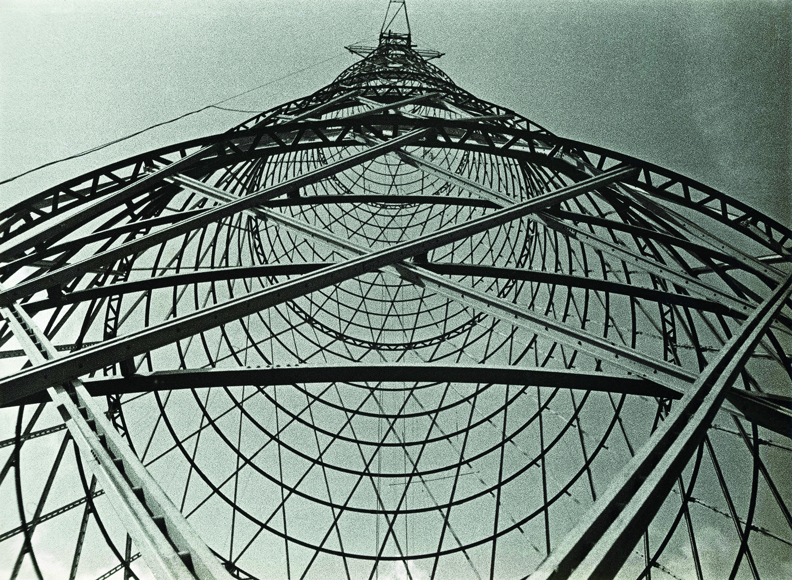 Fotografia em preto e branco. Vista de baixo de uma torre de transmissão, que destaca as linhas diagonais e paralelas e os arcos que formam a estrutura de aço da torre.