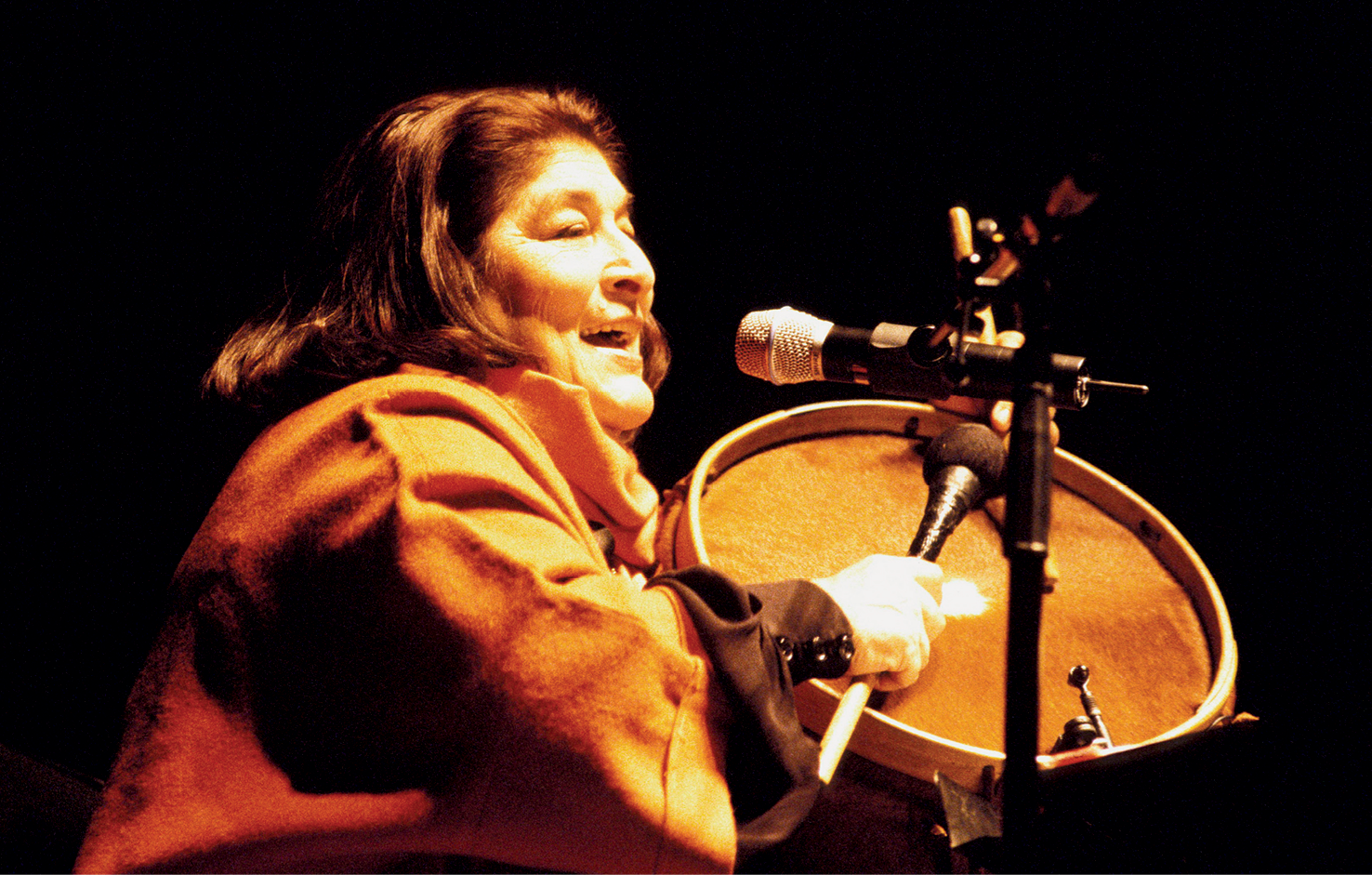 Fotografia. Uma mulher com cabelos na altura dos ombros, usando um casaco. Ela está sentada, com a boca aberta perto de um microfone e segurando com uma das mãos um bumbo e, com a outra, uma baqueta.