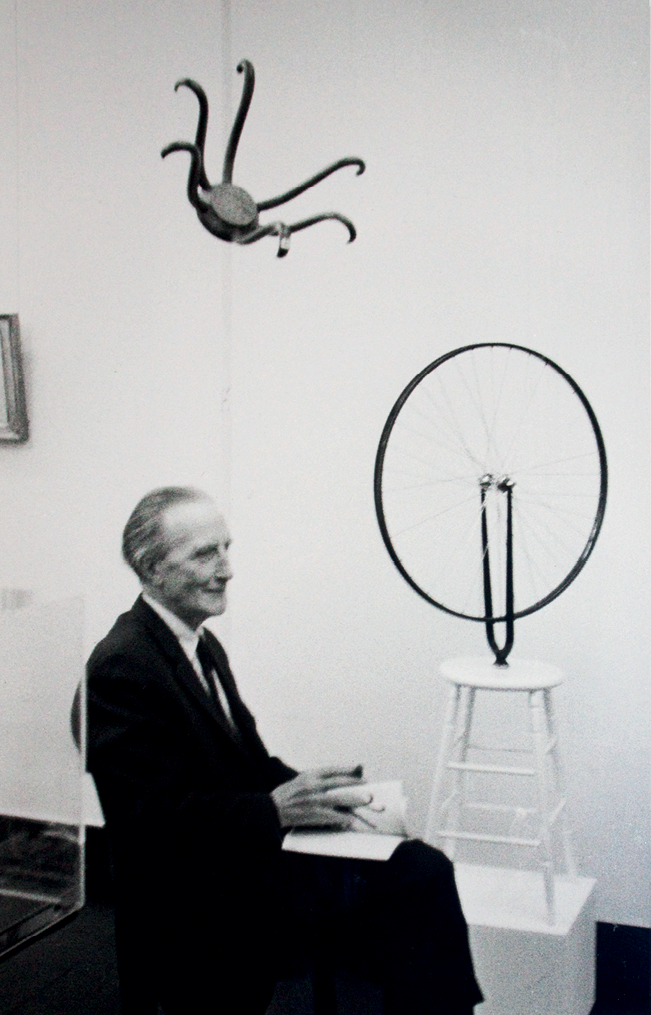 Fotografia em preto e branco. Homem com cabelos grisalhos, usando terno e gravata, está sentado em uma cadeira de perfil. Ao lado dele, há obras de arte.