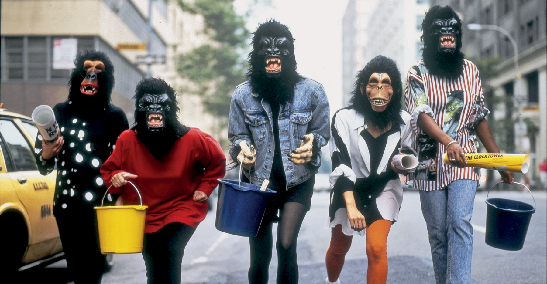 Fotografia. Cinco mulheres usando roupas coloridas e máscara de gorilas. Elas estão segurando baldes e rolos de papel, e andam no meio da rua. Ao redor, há prédios e um táxi.