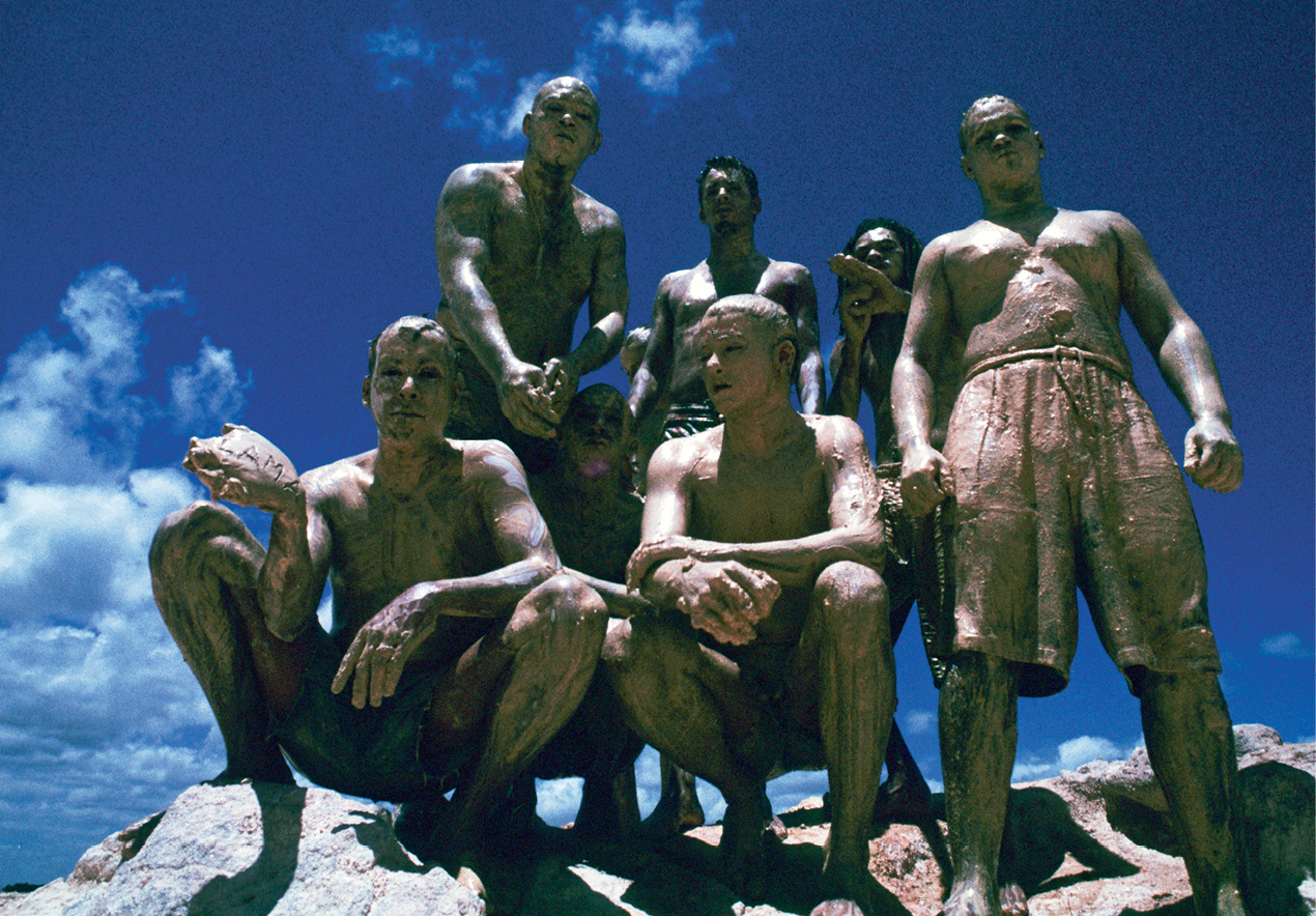 Fotografia. Sete homens, três agachados e quatro em pé, sobre uma pedra. Eles vestem apenas bermudas e seus corpos estão cobertos de lama. Ao fundo, um céu azul com algumas nuvens.