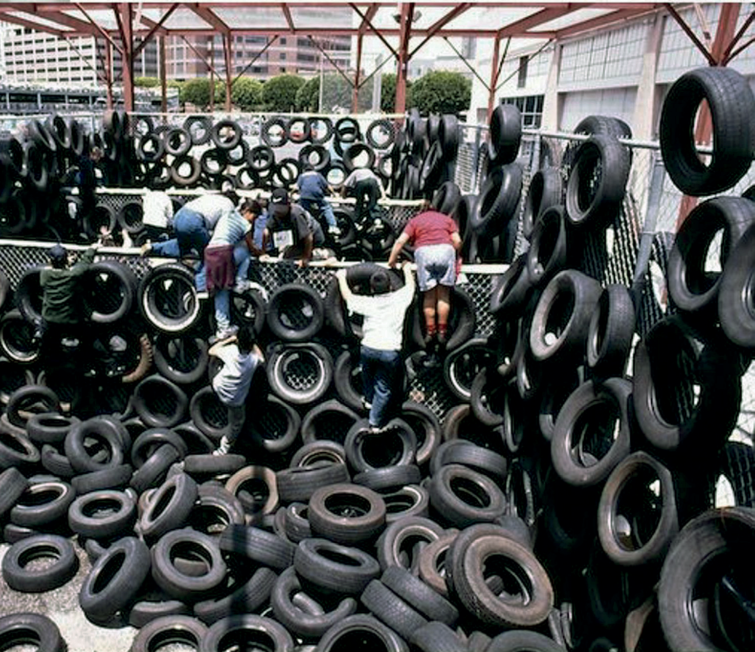 Fotografia. Espaço delimitado por alambrados de metal, com vários pneus pendurados e empilhados no chão. Diversas pessoas estão escalando os pneus e saltando os alambrados.