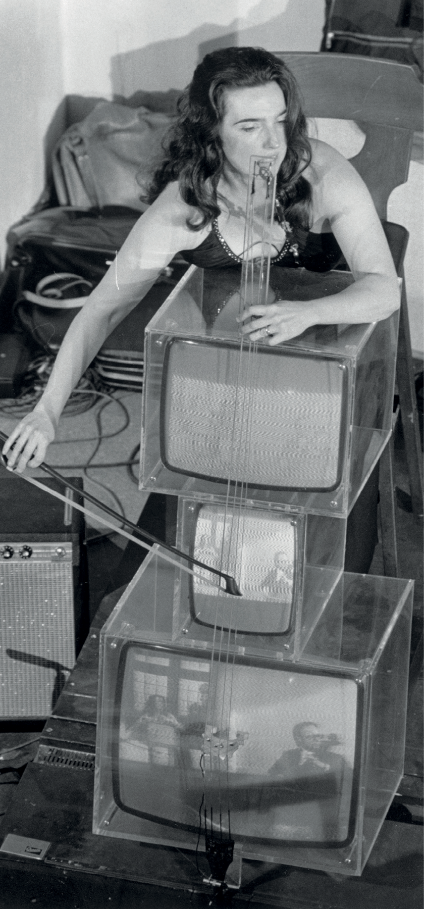 Fotografia em preto e branco. Uma mulher com cabelos escuros e compridos, usando um vestido, está de pé e toca um violoncelo que é feito de três televisores de tubo empilhados. Os televisores têm estrutura de acrílico.