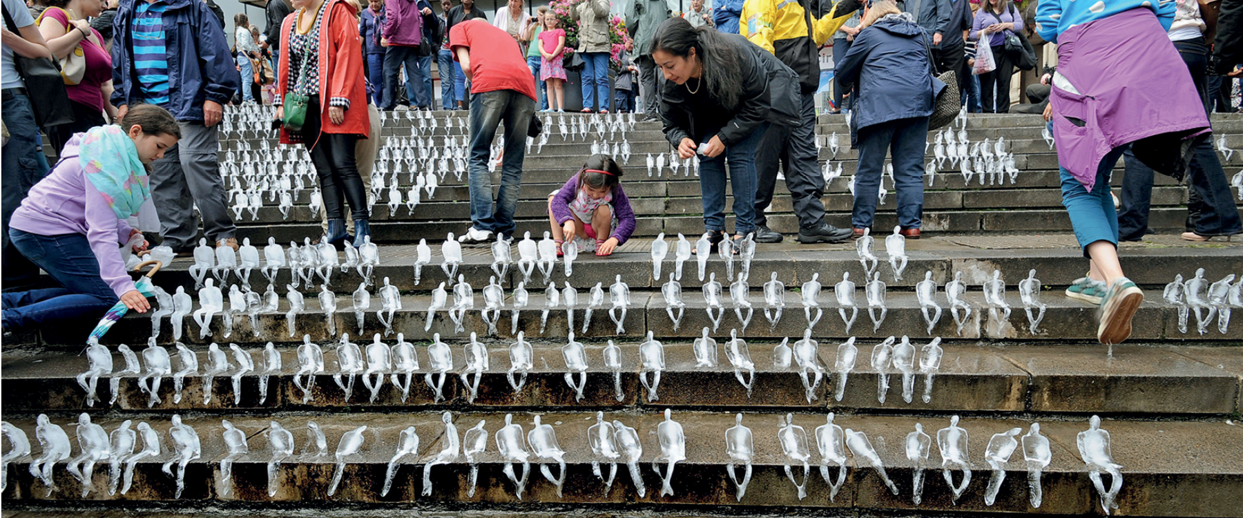 Fotografia. Nos degraus de uma escadaria há centenas de esculturas de gelo com formato de pessoas em miniatura sentadas de pernas cruzadas. Em volta das esculturas, há crianças e adultos que as observam.