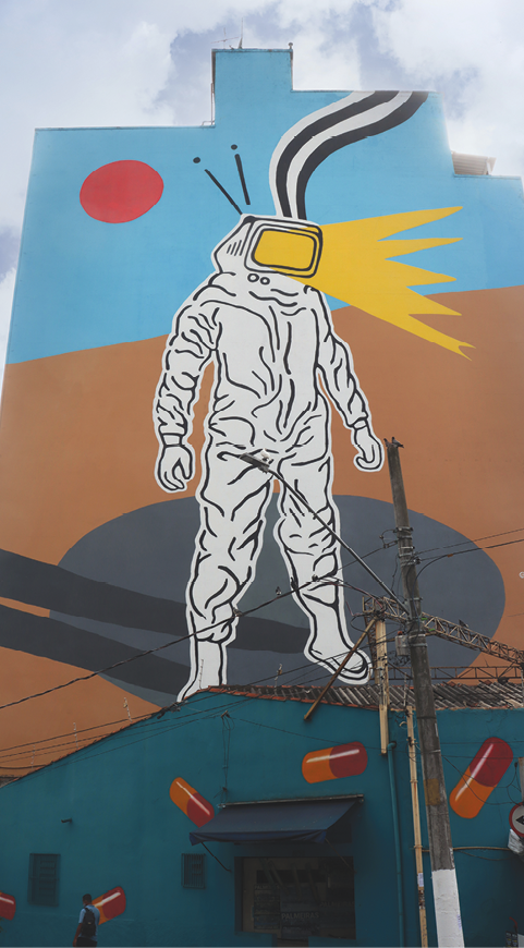 Fotografia. Grafite na fachada de um prédio, com a figura de um astronauta; do visor do capacete dele sai uma luz amarela; ao fundo há um círculo vermelho em um céu azul, e sob os pés do astronauta, a sombra dele projetada no chão marrom e um círculo cinza.