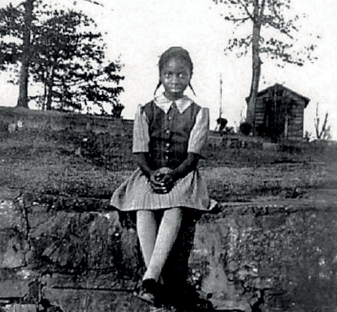 Fotografia em preto e branco. Uma menina negra com cabelos presos em duas tranças laterais, usando vestido com botões. Ela está sentada em uma mureta com as mãos juntas sobre o colo. Ao fundo, há um muro, árvores e uma pequena casa.