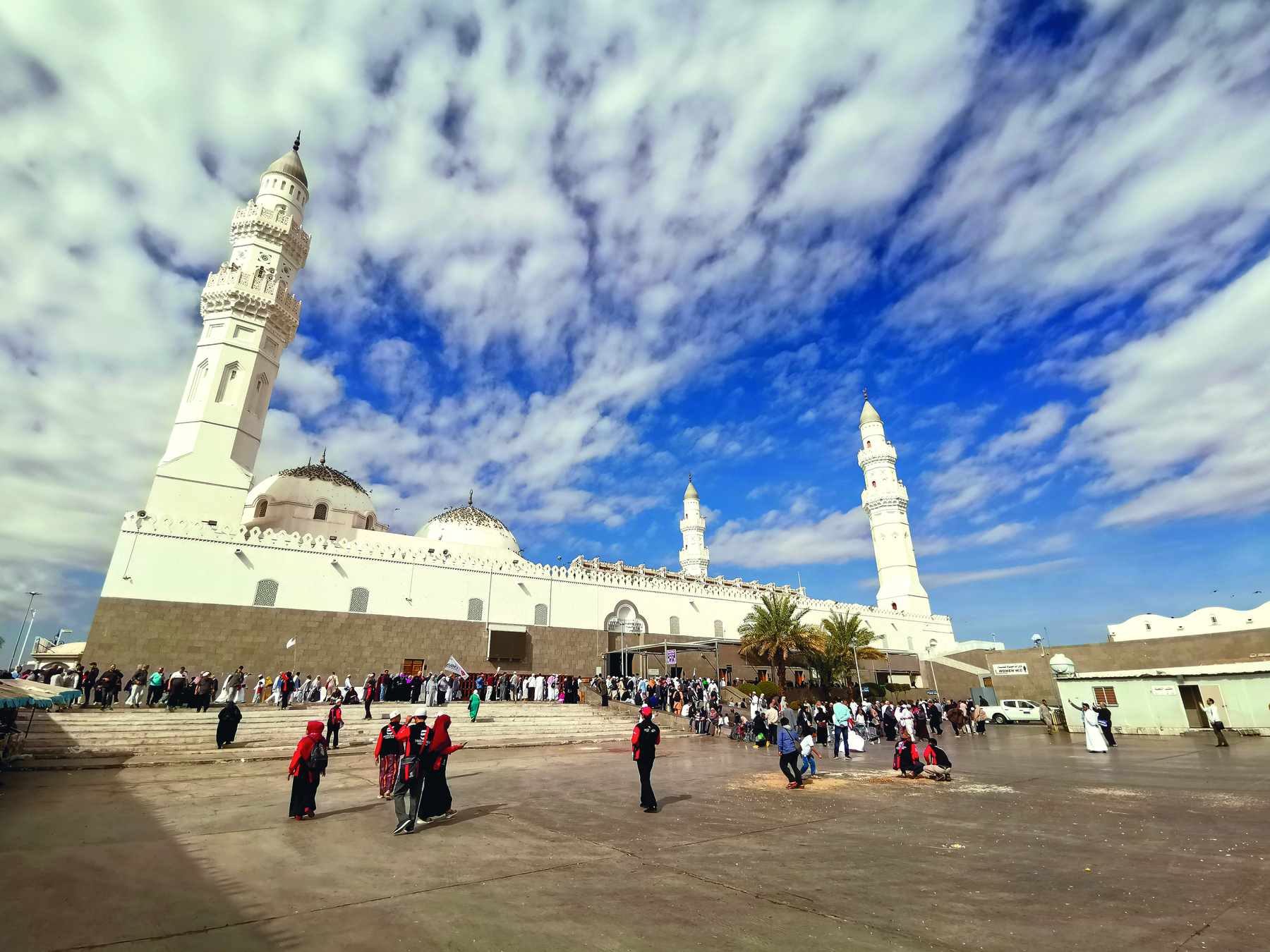 Fotografia. Pessoas em frente a uma grande mesquita de muros brancos. Nas extremidades dos muros, grandes torres chamadas minaretes. No centro do muro uma porta.