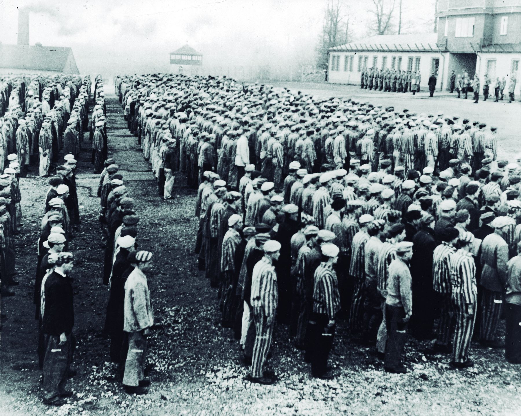 Fotografia em preto e branco. Homens enfileirados em um campo de concentração. Eles usam roupas largas e um gorro na cabeça. A roupa de alguns é listrada. Ao fundo, soldados perfilados e algumas edificações.