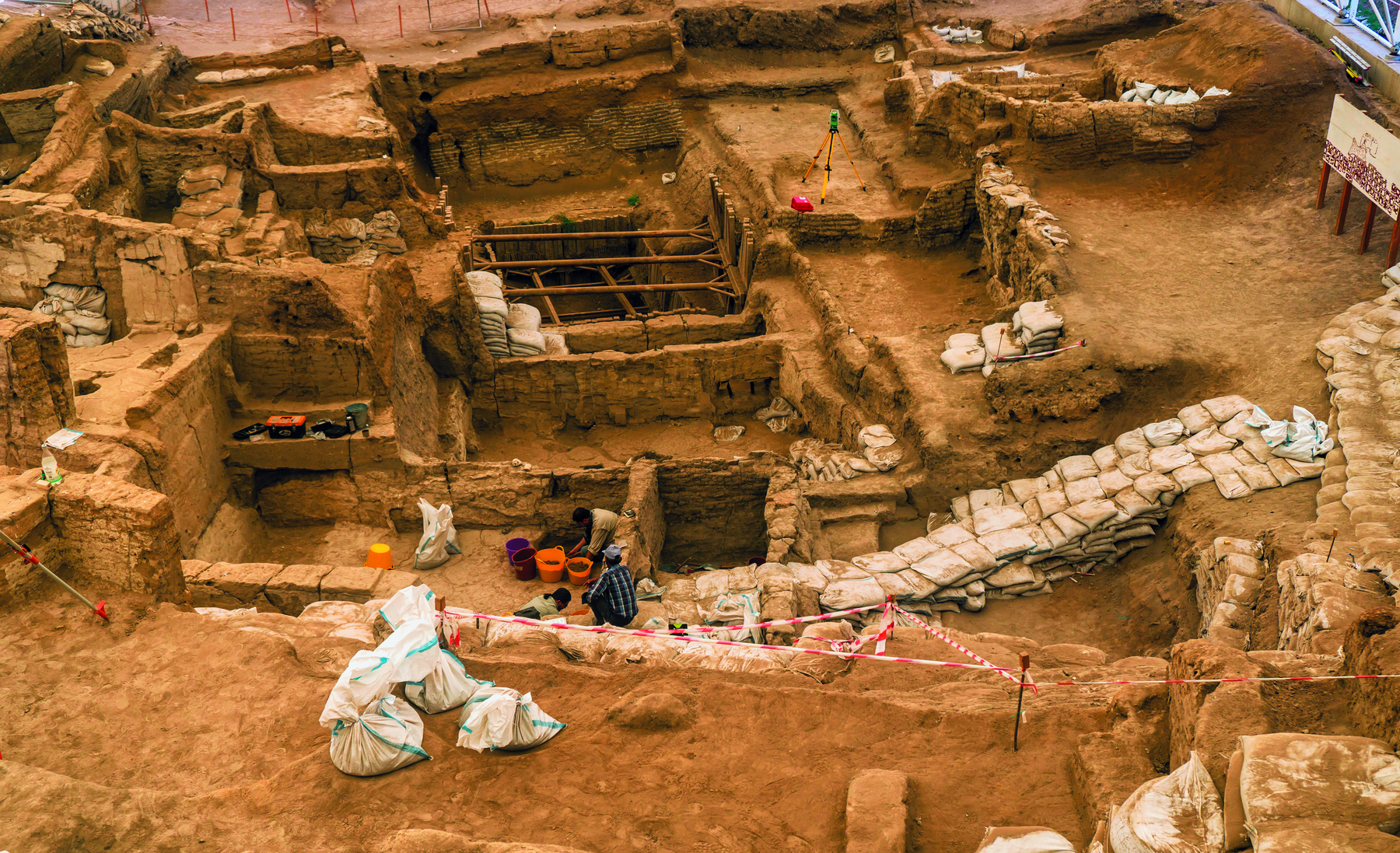Fotografia. Vista aérea de arqueólogos trabalhando em um solo terroso. No terreno escavado, há paredes e muros de pedra, indicando ambientes separados de tamanhos variados, em vários níveis diferentes. Sobre o solo, diversos sacos com terra.
