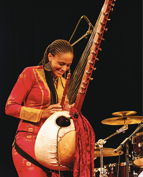 Fotografia. Mulher negra com os cabelos trançados e roupa vermelha. Ela segura um instrumento de base arredondada. Na parte superior, uma haste vertical e cordas, semelhante a um braço de violão, mas dispostos como em uma harpa.