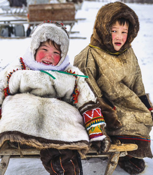 Fotografia. Duas crianças sentadas sobre um suporte de madeira na neve. Elas vestem roupas peludas e compridas, estão apenas com o rosto à mostra. Suas bochechas estão avermelhadas por causa do frio.