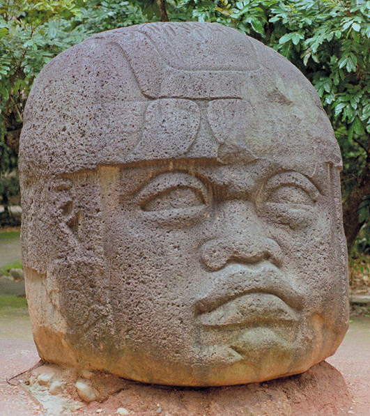 Fotografia. Cabeça gigante esculpida em uma pedra arredondada. O nariz é achatado, os lábios são largos, e os olhos amendoados.