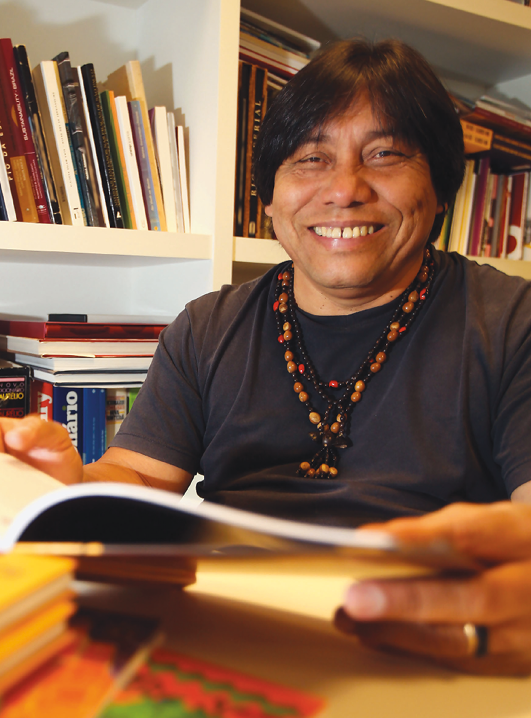 Fotografia. Destaque para um homem indígena sorridente, tem o rosto arredondado, cabelo liso e preto. Usa alguns colares. Está sentado à mesa com um livro nas mãos. Atrás dele, estante com muitos livros.
