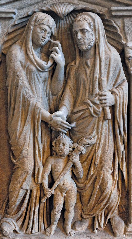 Escultura. Imagem esculpida em alto relevo. Casal de mãos dadas. Ambos usam longas túnicas. Entre eles, na frente uma criança pequena. Ela está nua e segura uma pequena tocha nas mãos.
